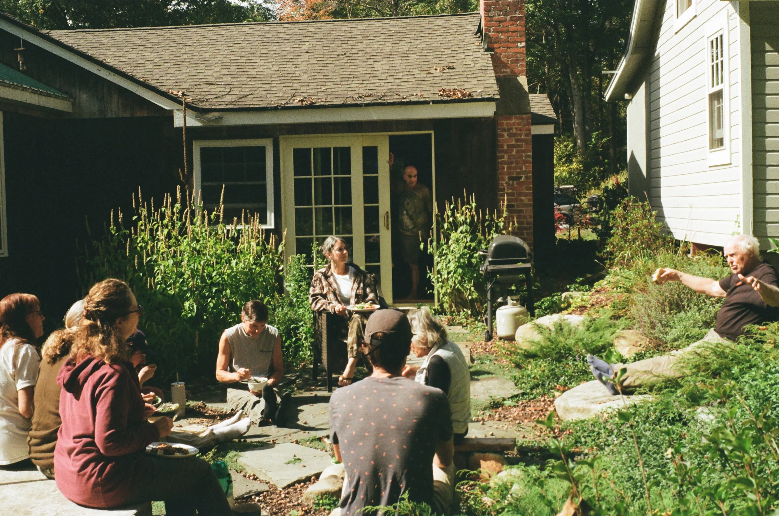A group of people gathered in a backyard for a social event on a sunny day. They are sitting on the ground and benches, eating and talking, with some standing or relaxing. There is a woman with a tray, and an elderly man gesturing with his arms. The 