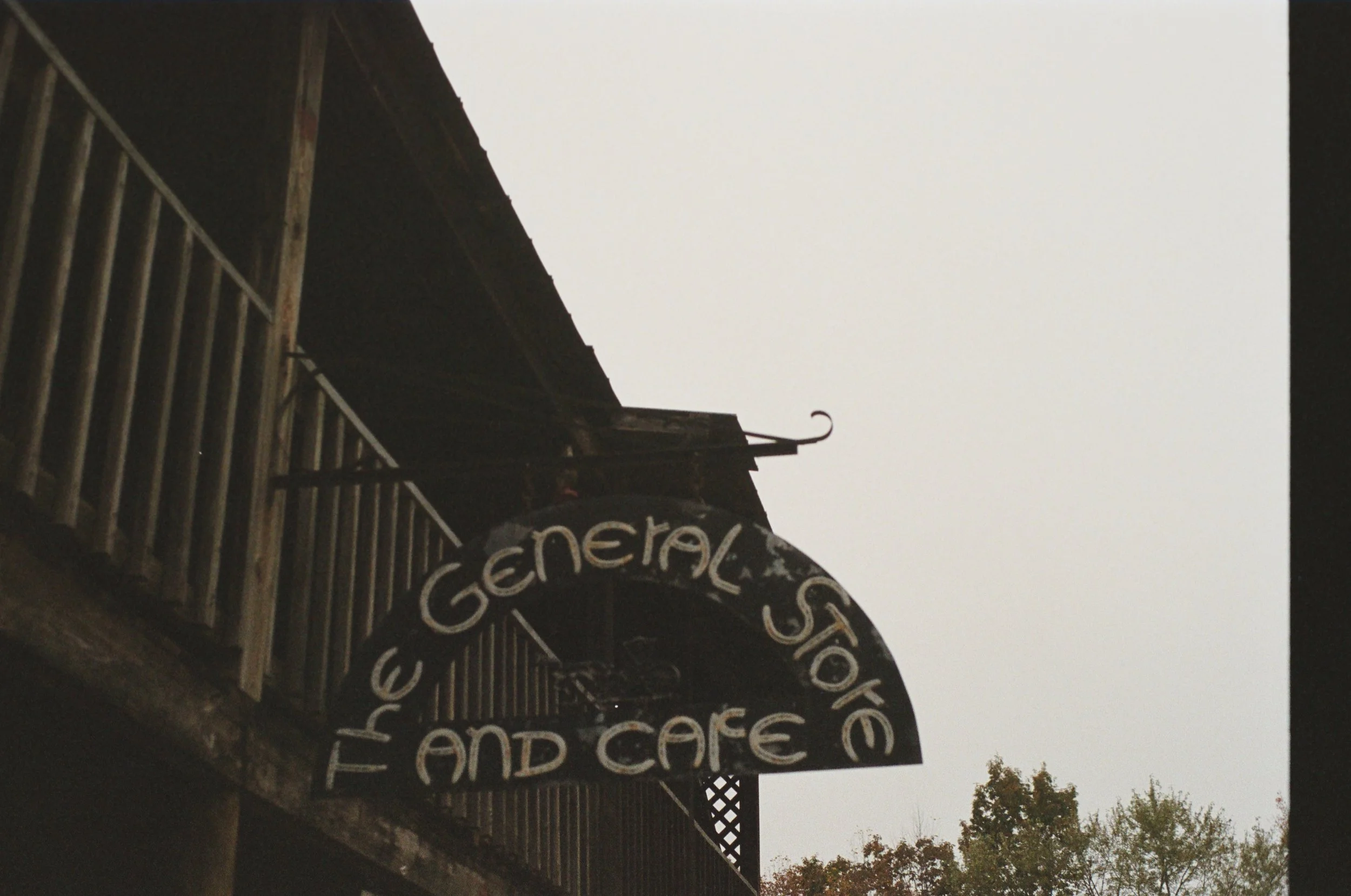 A dark curved sign reading 'The General Store and Cafe' hanging from a building with a wooden balcony, against a cloudy sky and some trees in the background.