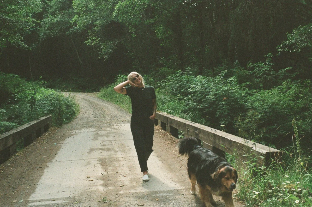 A woman walking her black and brown dog on a dirt trail through a forested area.
