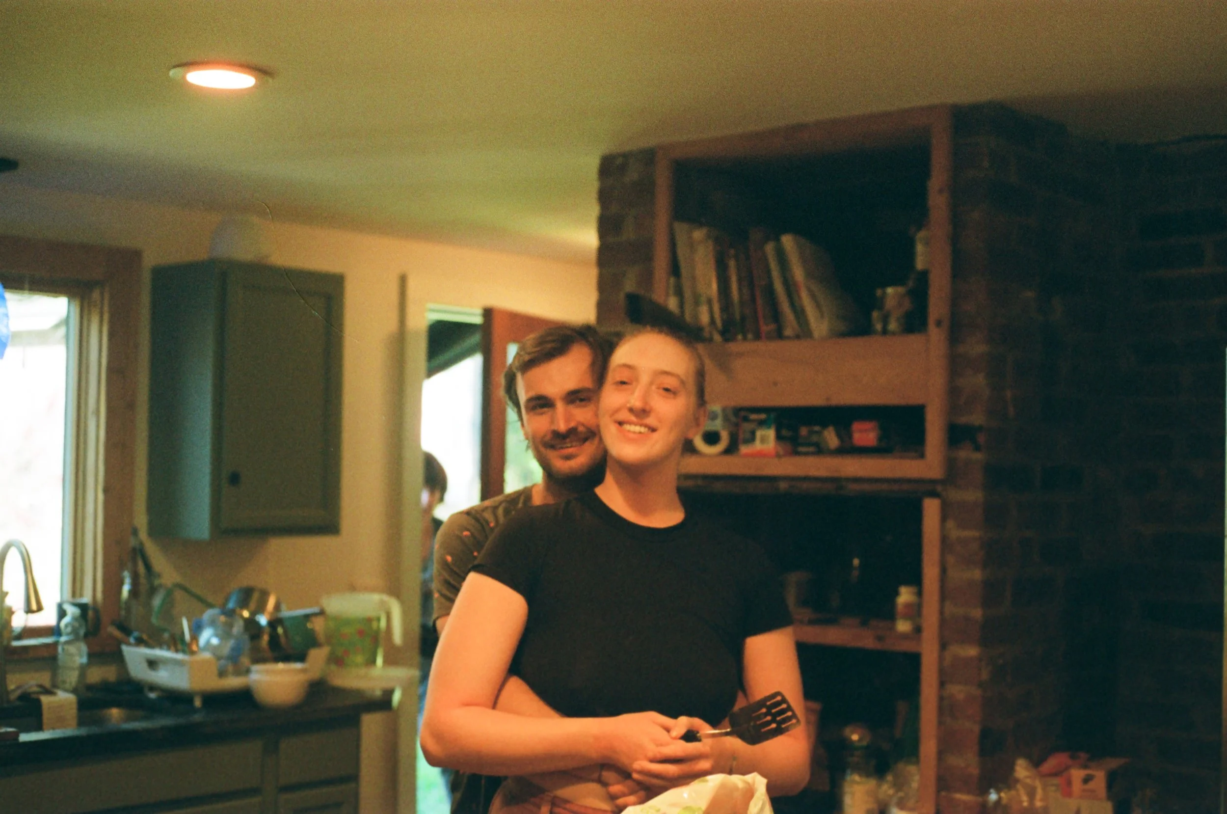 A smiling man and woman standing close inside a home, with the man behind the woman, both looking at the camera. The woman is holding a spatula.