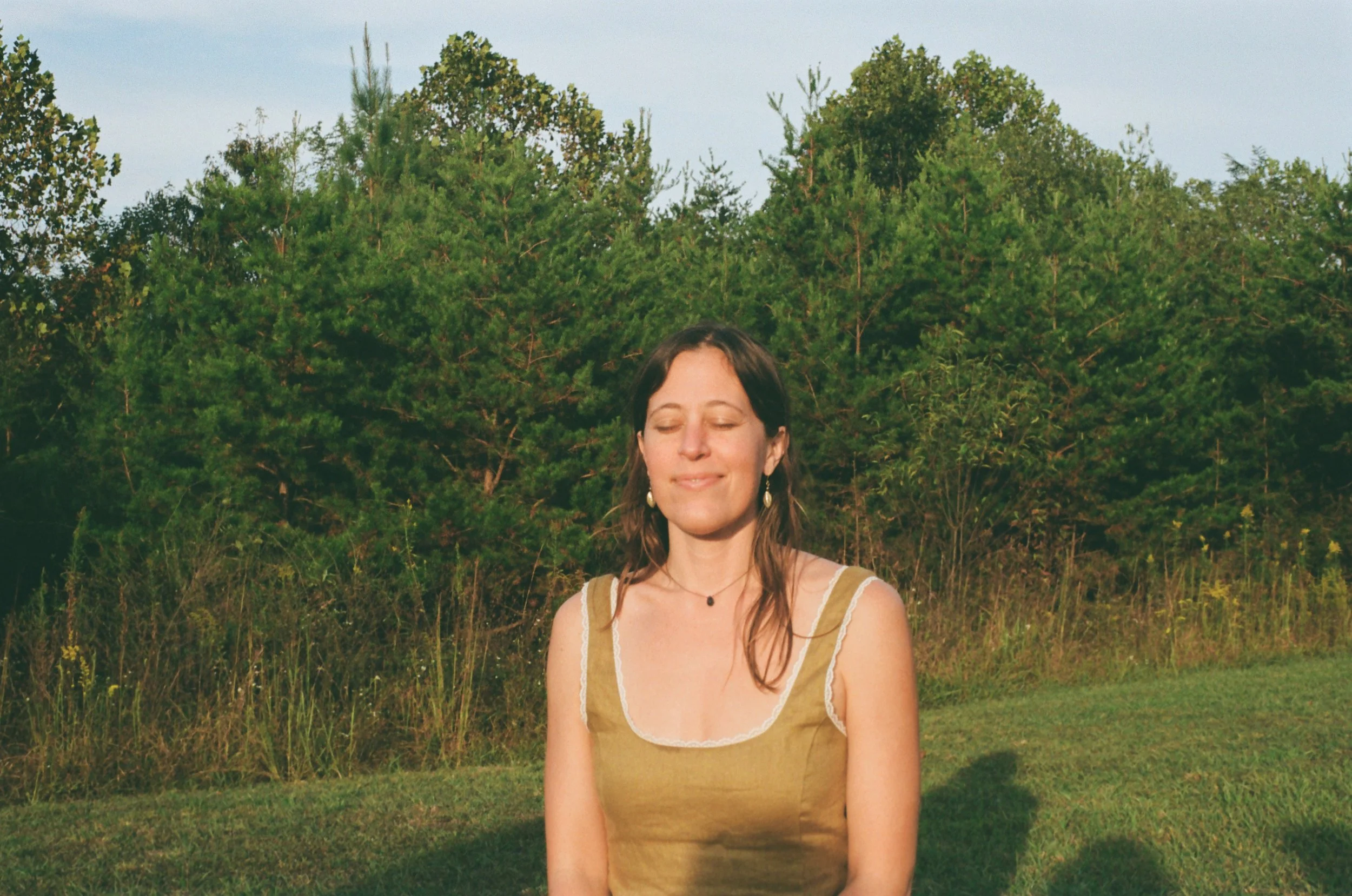 A woman with long brown hair, wearing a sleeveless yellow top with white trim, standing outdoors with her eyes closed and a calm expression, against a background of green trees and grass.
