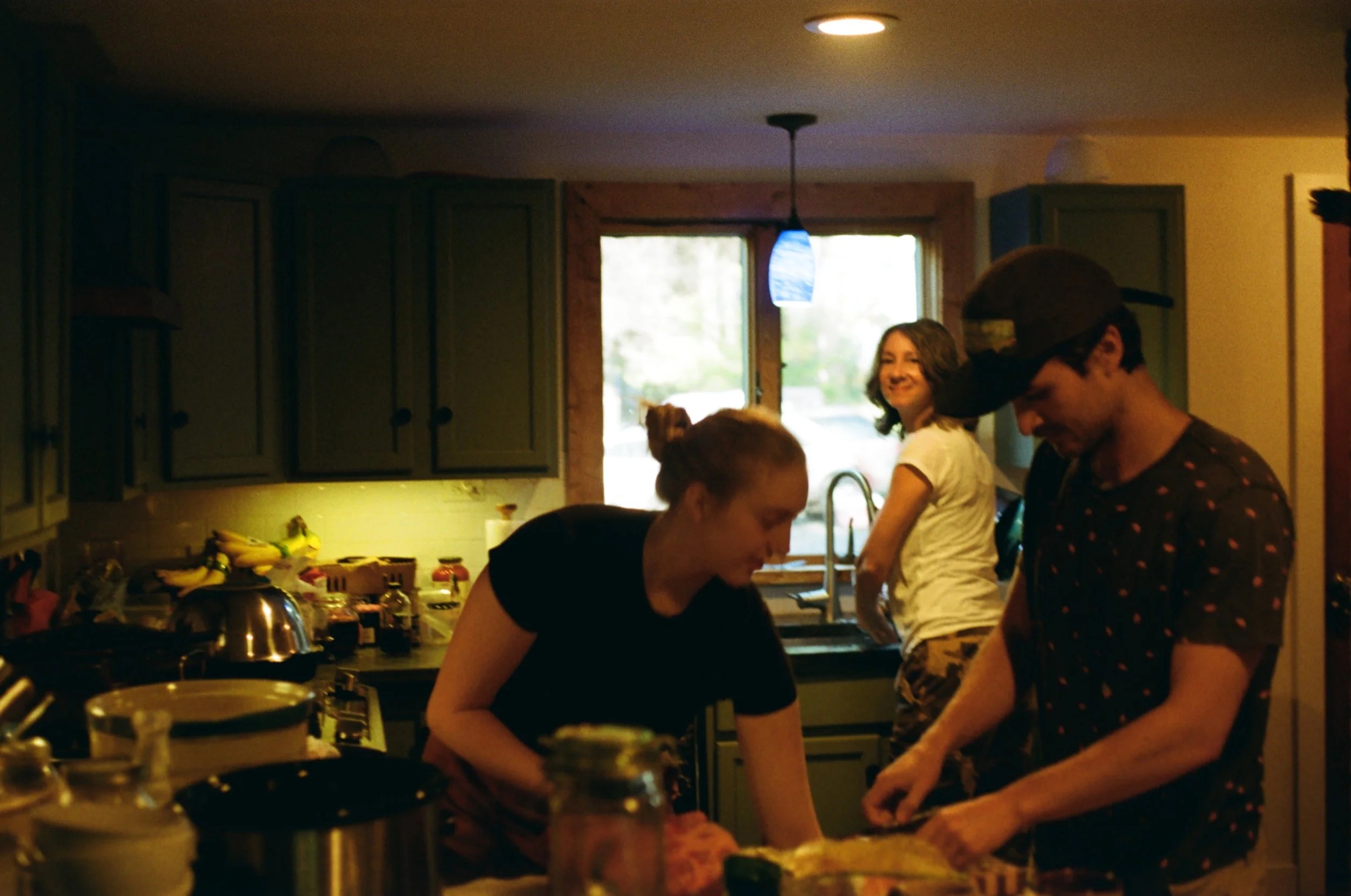 A group of young people in a kitchen, some preparing food and smiling, with a window in the background letting in natural light.