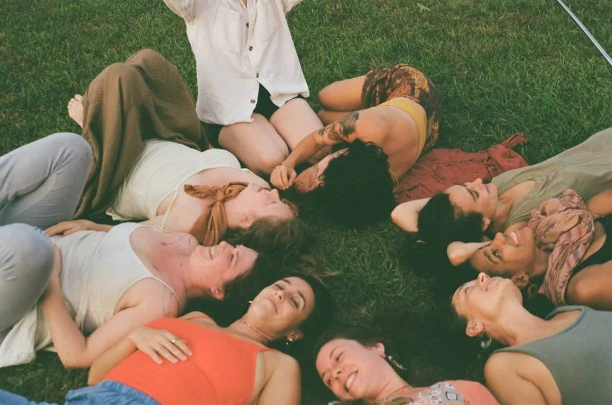 A group of women lying on the grass, forming a circle with their heads close together, smiling and enjoying each other's company outdoors.