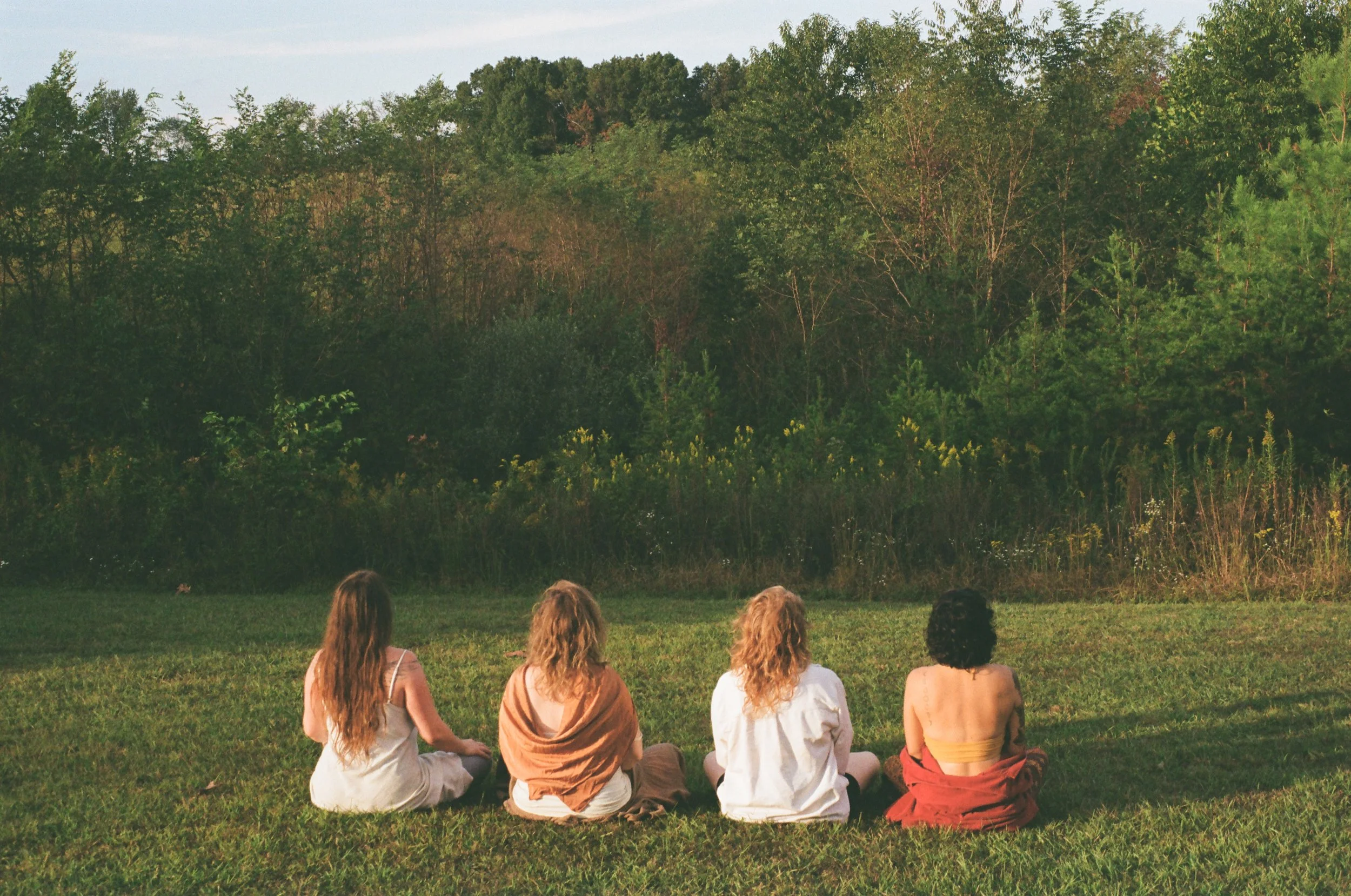 Four women sitting on the grass facing a forested area, with trees and bushes in the background, during daytime.