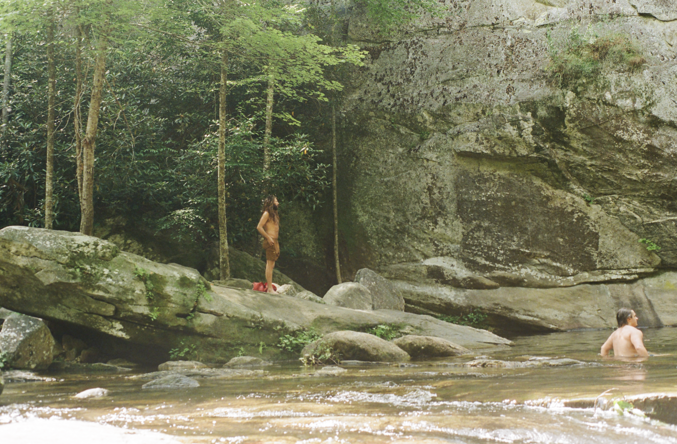 A man and a woman in a forested river with large rocks and a mossy cliff.