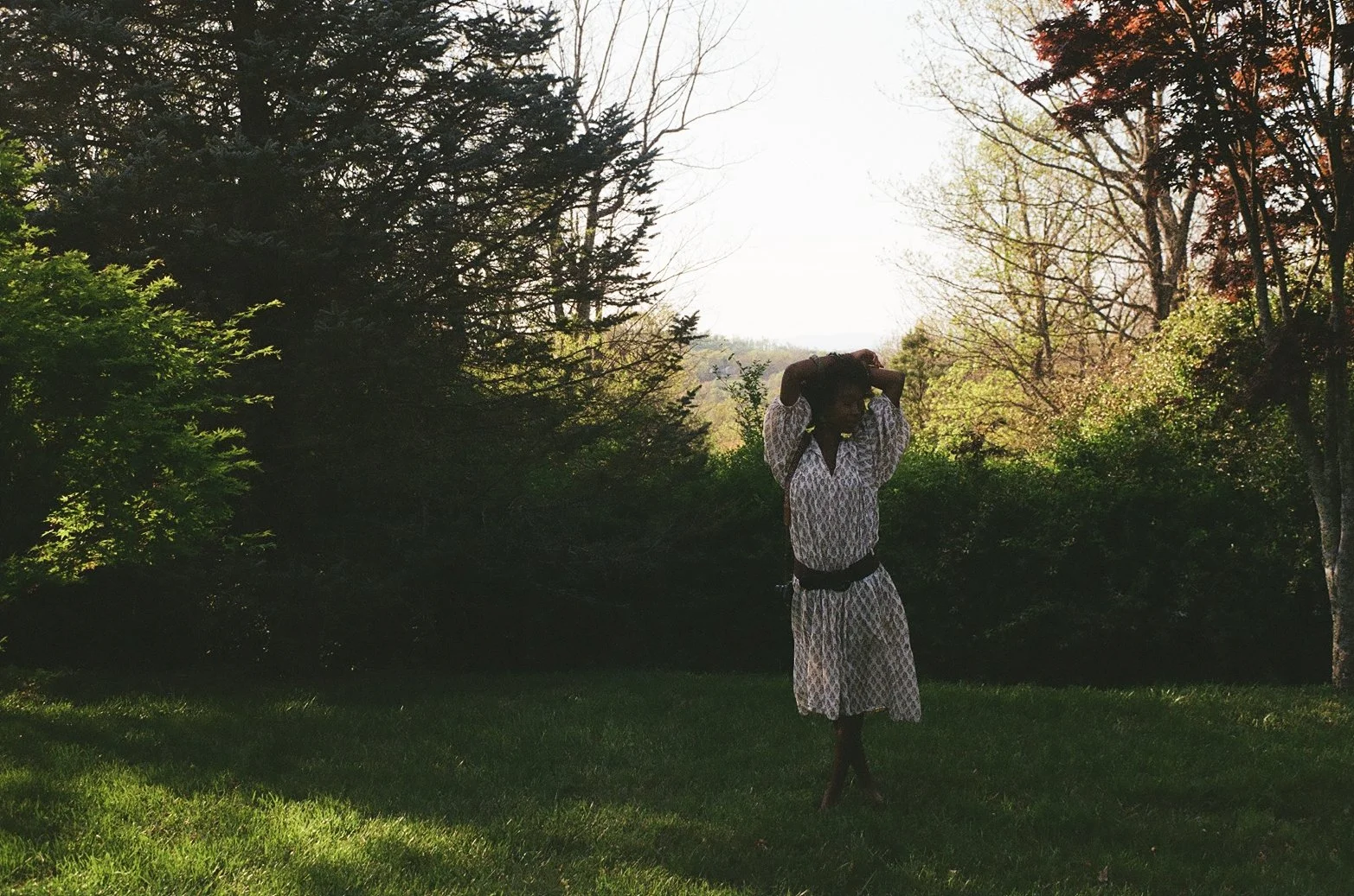 A woman in patterned dress standing on grass with trees and bushes around, backlit by bright sunlight.