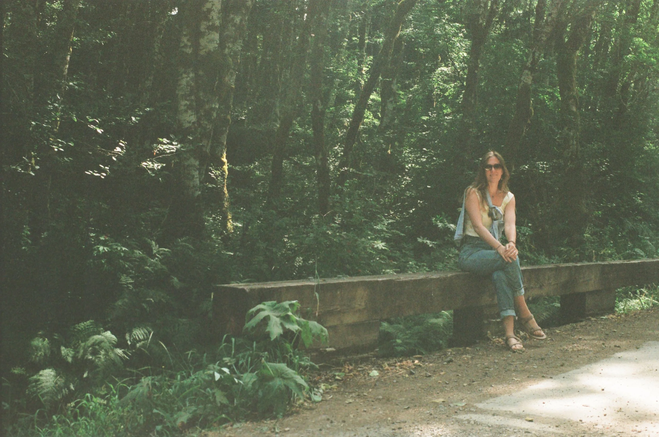 A woman sitting on a wooden bench in a forested area, surrounded by lush green trees and plants, with sunlight filtering through the leaves.