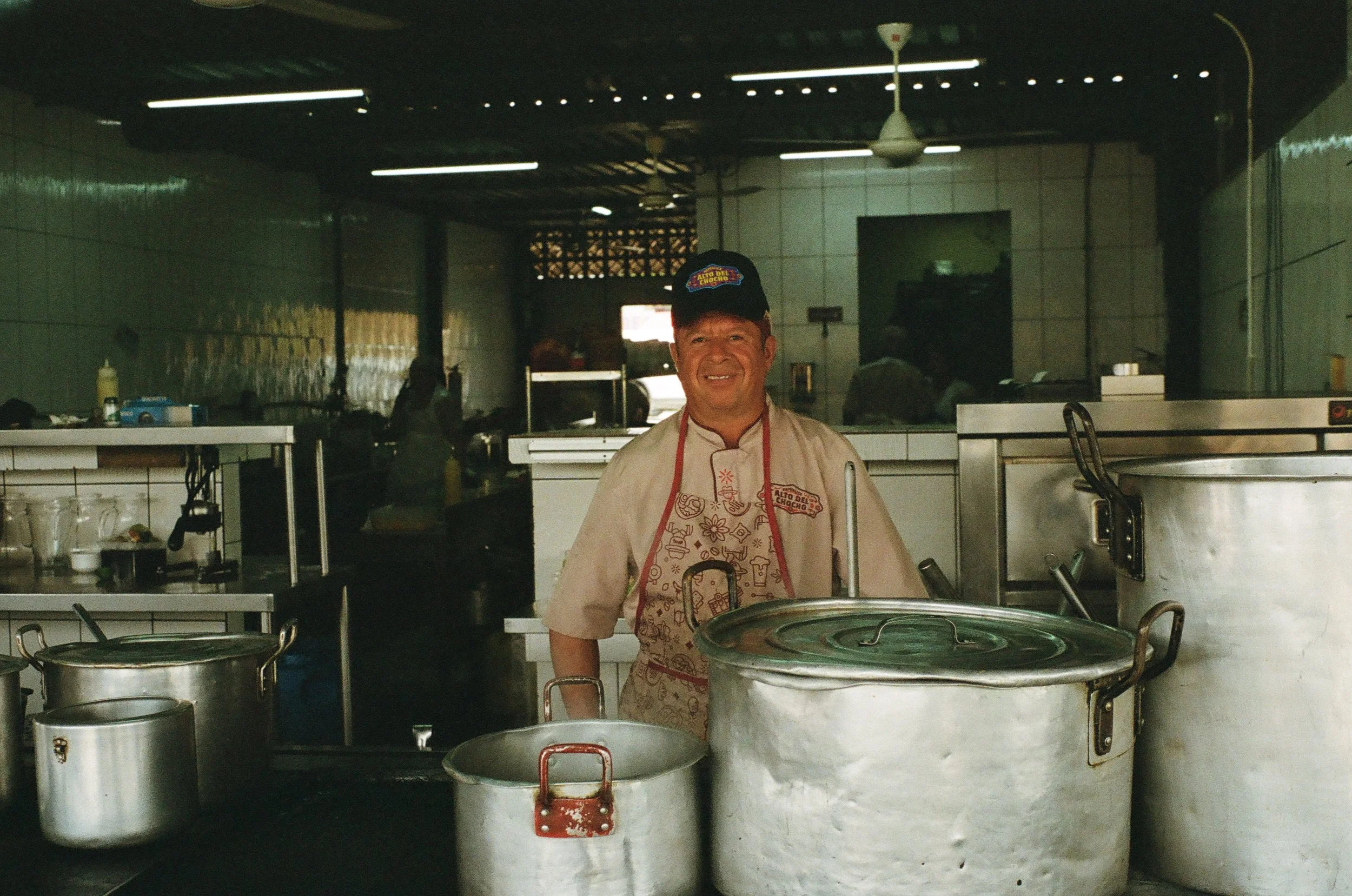 A smiling man in a chef's uniform and hat stands behind large metal cooking pots in a kitchen.