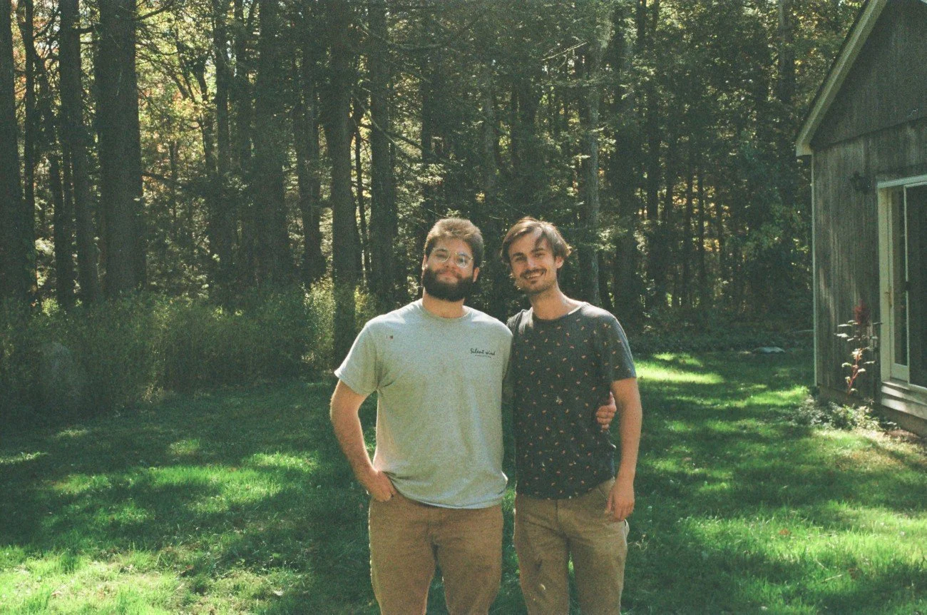 Two young men standing close together outdoors in a backyard, smiling. One has a beard and glasses, wearing a light t-shirt, and the other has short hair, wearing a dark shirt with a pattern. Trees and a wooden house are in the background.