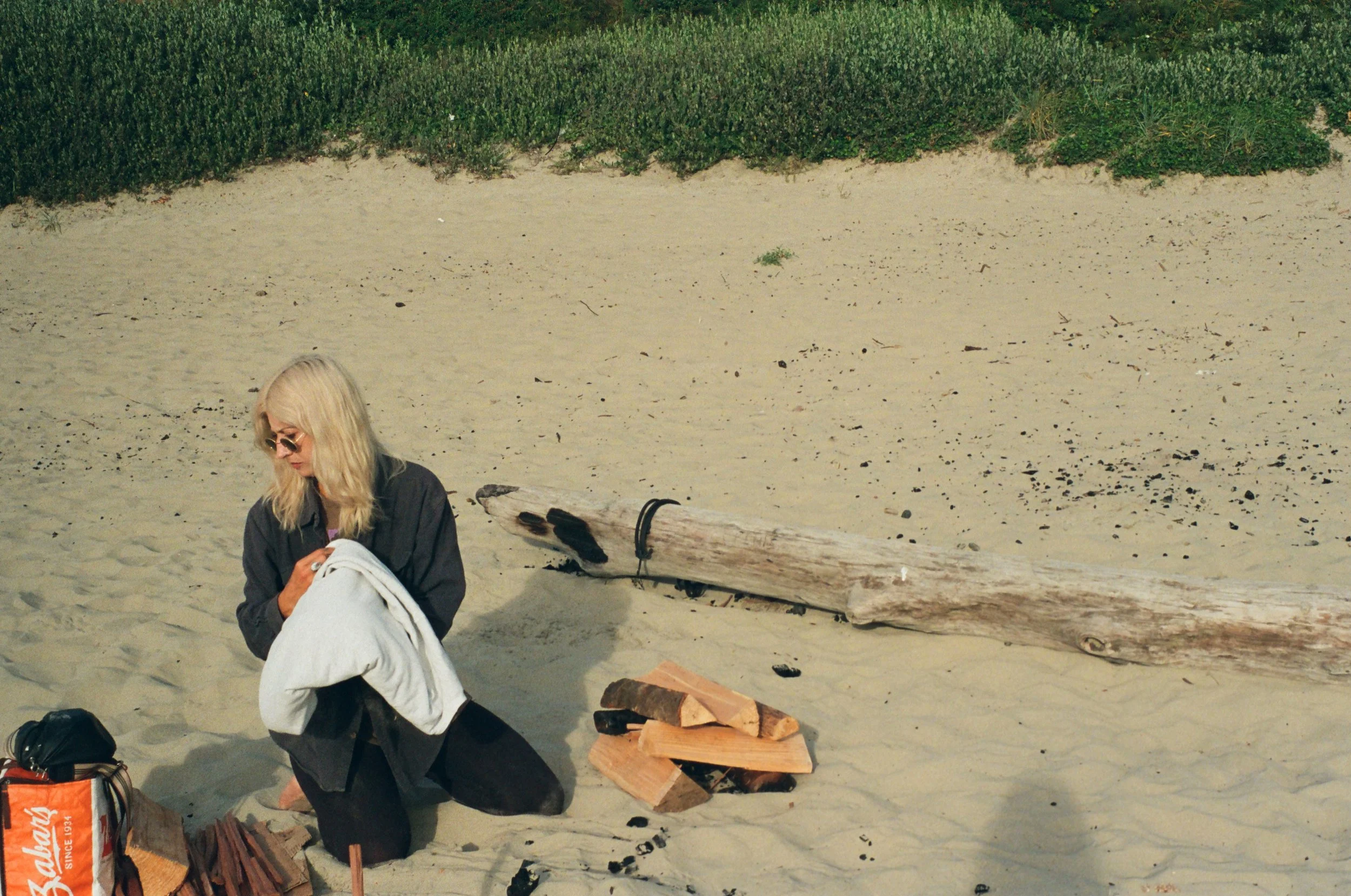A woman with blonde hair, wearing sunglasses and a black jacket, kneeling on a sandy beach, holding a white towel or clothing item, with wood and logs nearby.