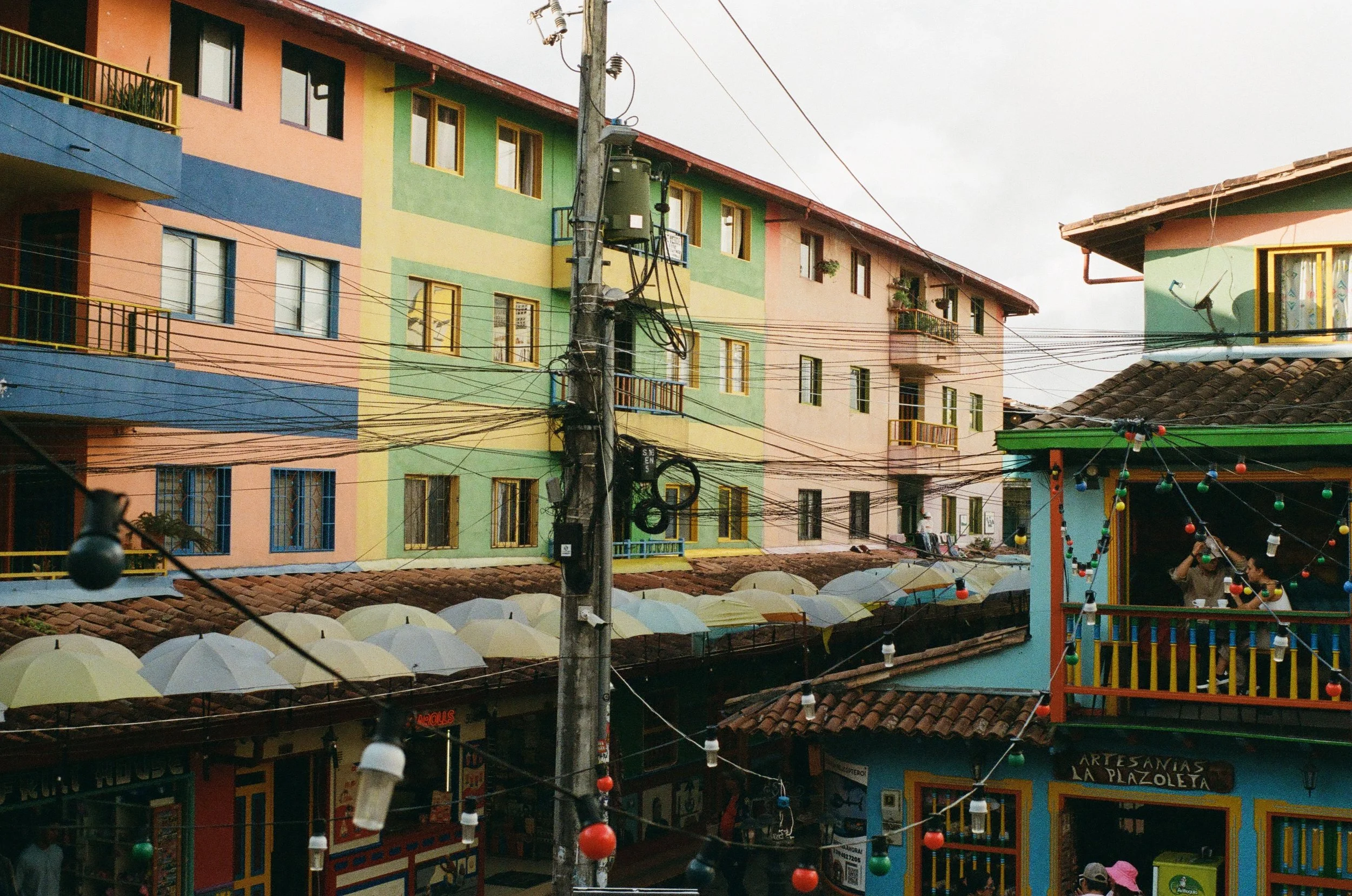 Colorful multi-story buildings with small balconies, laundry hanging, and decorative umbrellas on the roof, with string lights hanging across the street.