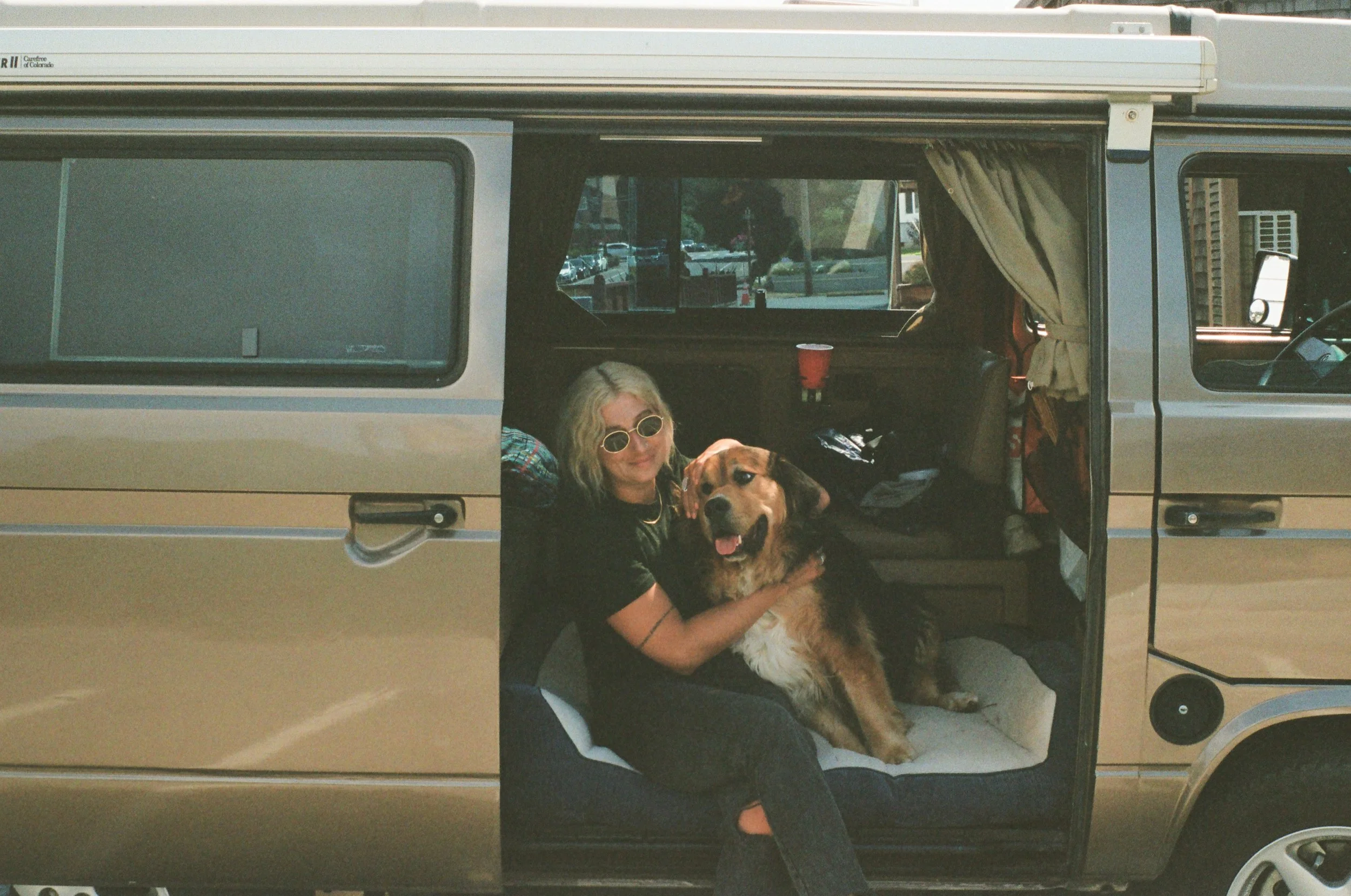 A woman with blonde hair and sunglasses sitting inside a vintage camper van, holding and cuddling a large golden retriever dog, with camping supplies and curtains visible inside.