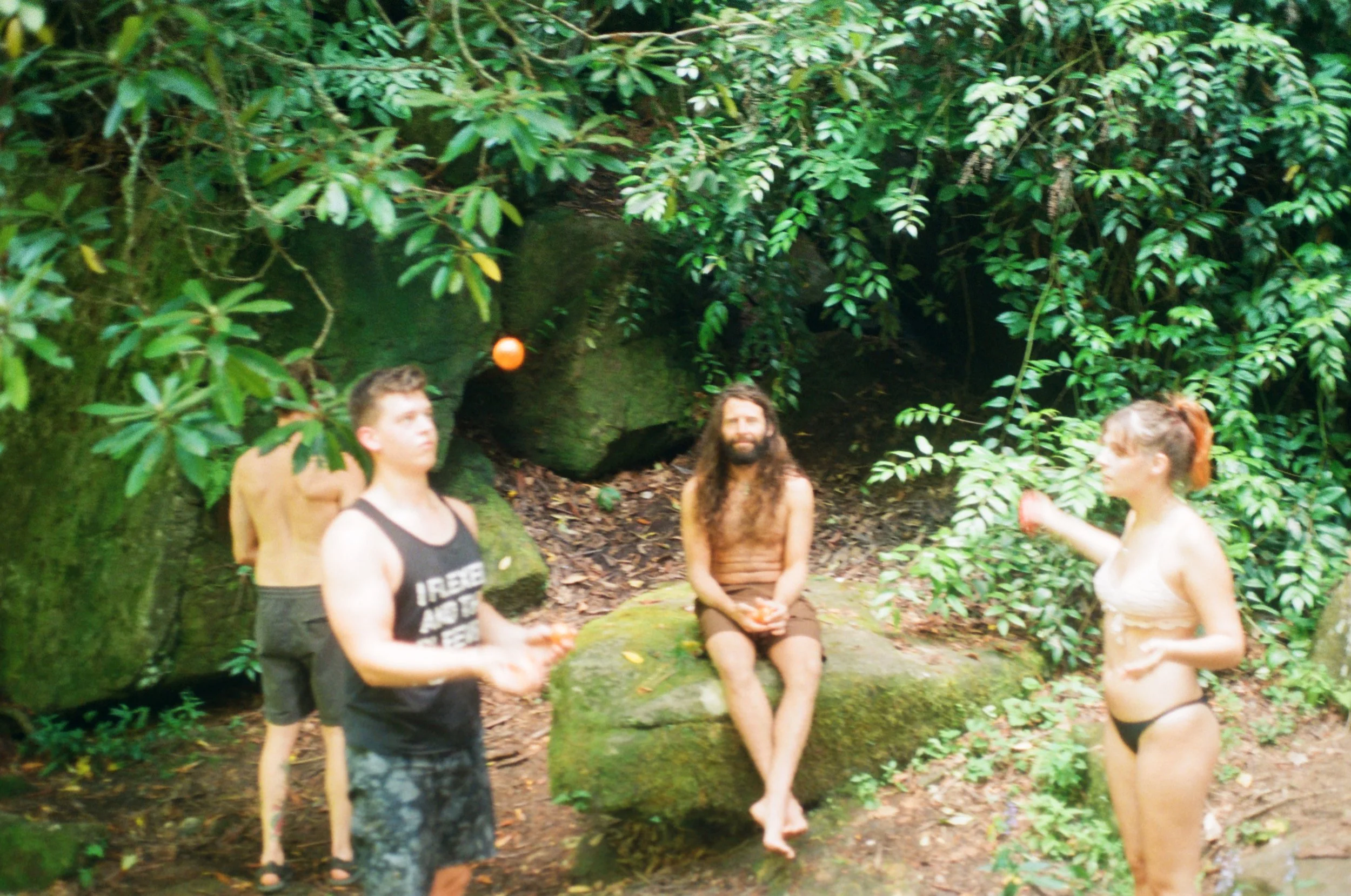 People playing catch with an orange ball in a forest setting, with lush greenery and rocks.