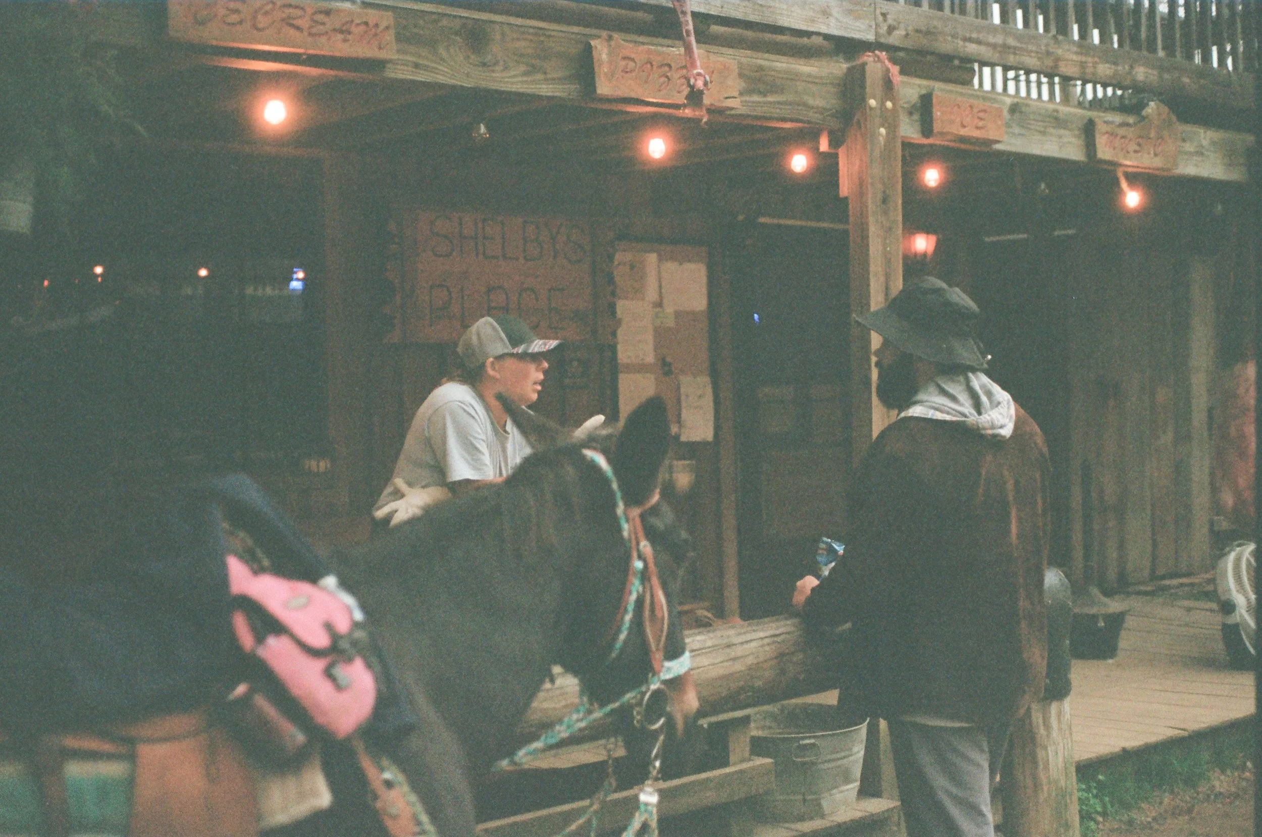 Two people conversing at Shelby's Place, a rustic wooden stand with string lights, in a casual outdoor setting. One person is leaning on a horse.