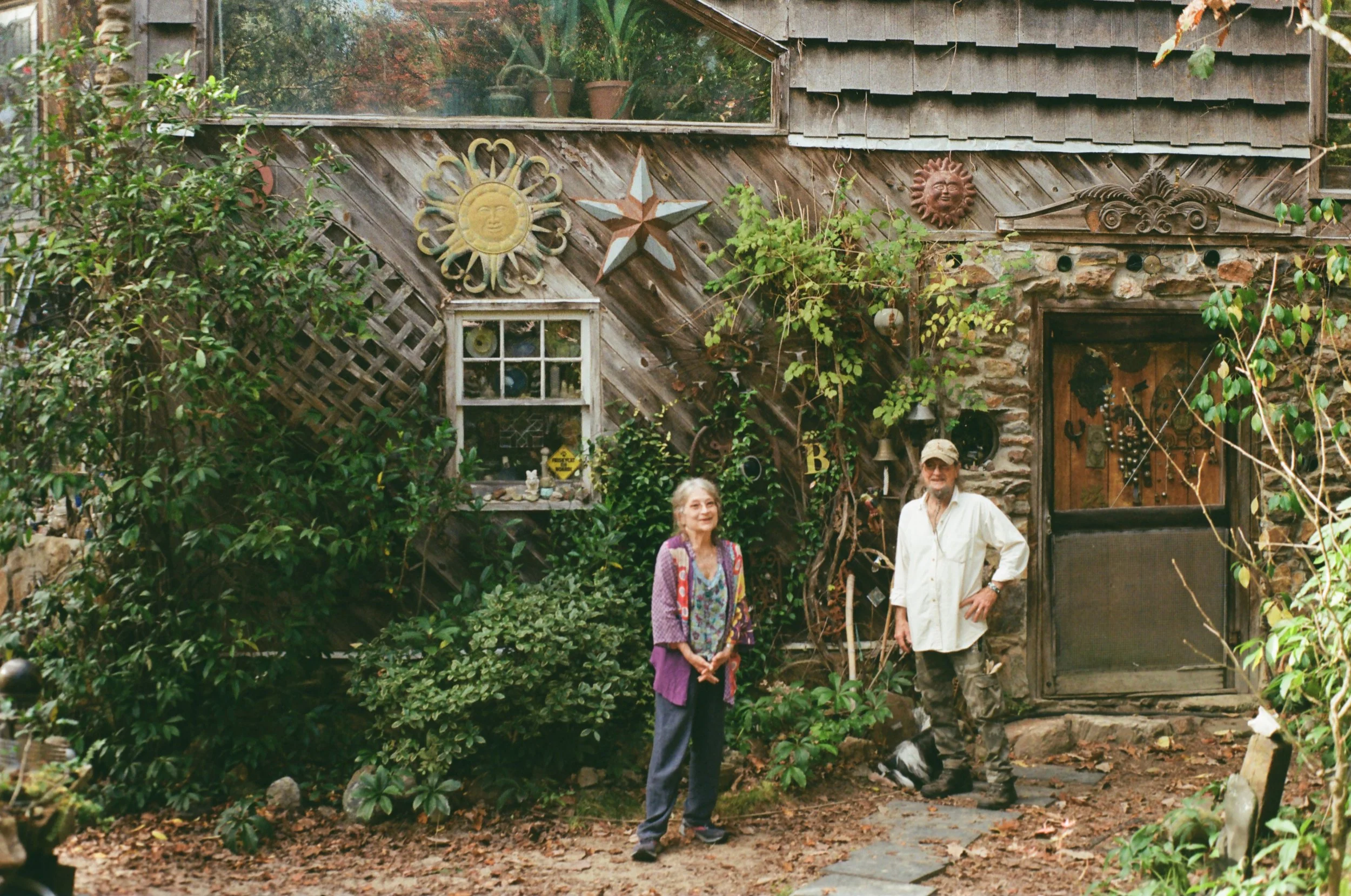 An elderly woman and an elderly man stand outside a decorated rustic house surrounded by plants and garden decorations. The woman wears a colorful patterned jacket, and the man wears a beige shirt and camouflage pants. The house has a stone and wood exterior with various ornaments, including a sun face and star, on the wall.