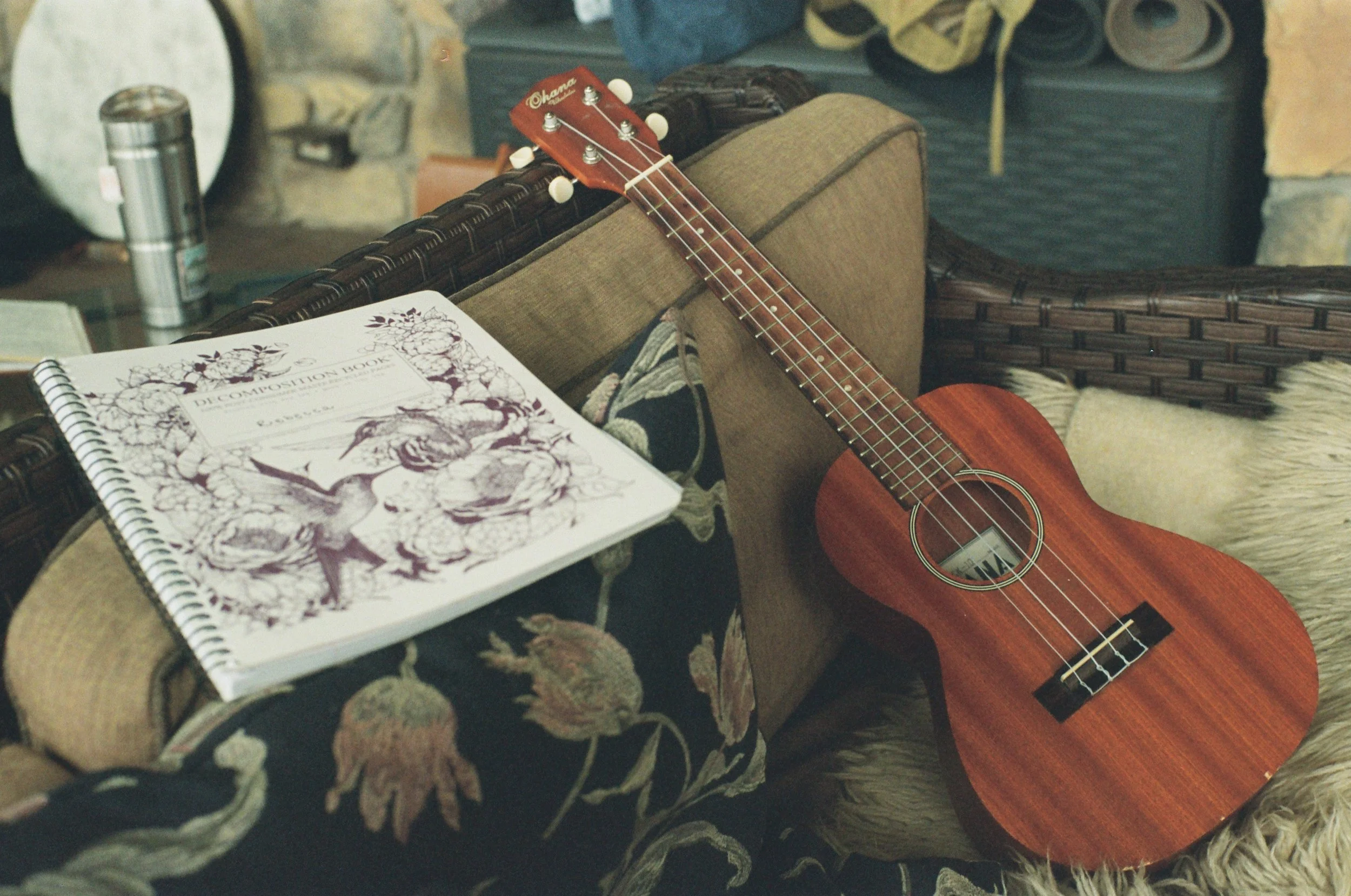 A small wooden ukulele with a red-brown finish resting on a wicker sofa with floral cushions, next to a spiral-bound music composition book on the sofa arm.