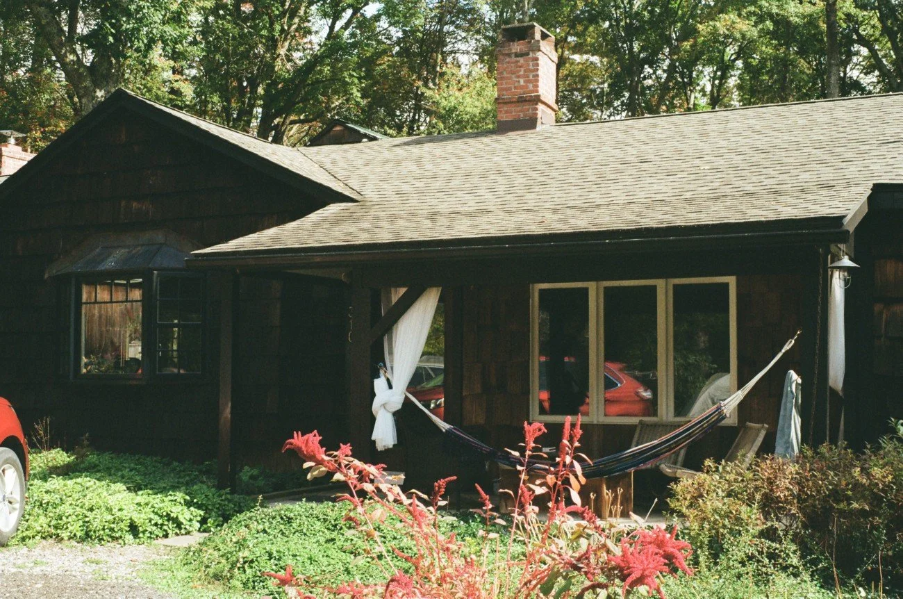 A rustic wooden house with a sloped roof and windows, a red car parked outside, a hammock tied between two posts, and lush greenery surrounding the house.