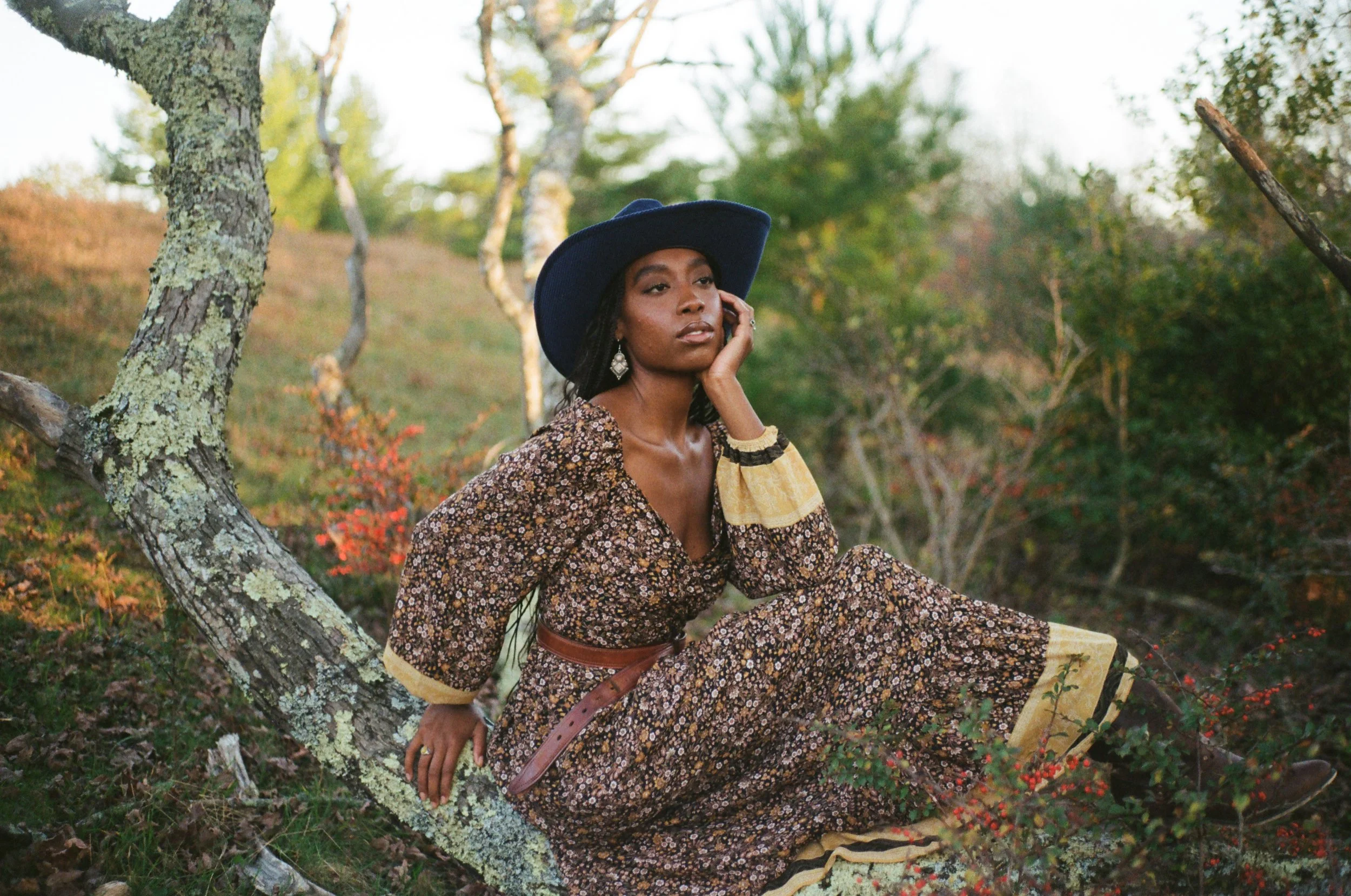 Woman sitting outdoors on a moss-covered tree branch, wearing a floral dress, a wide-brimmed hat, and jewelry, with a background of trees and greenery.