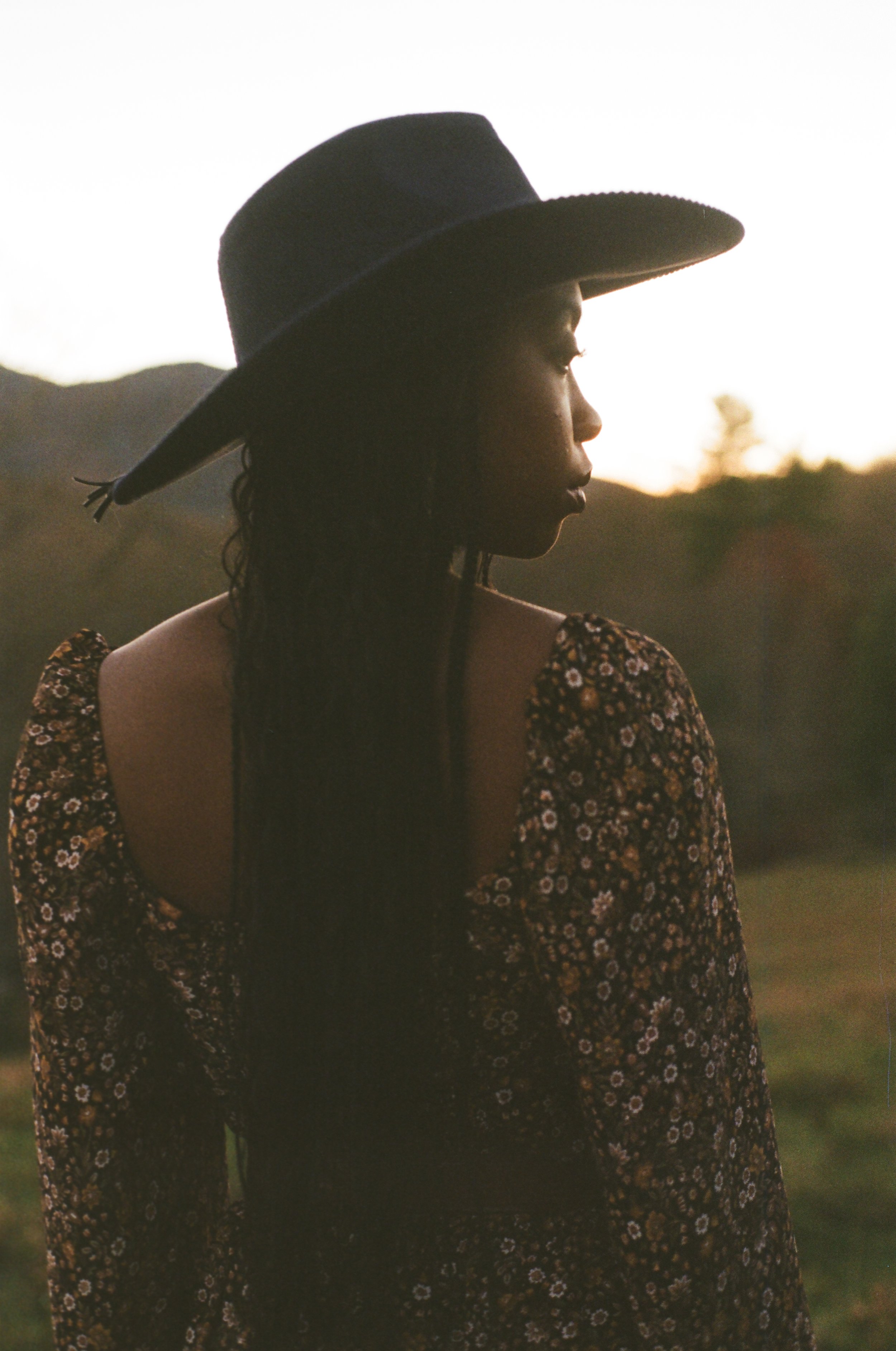 Side profile of a woman wearing a wide-brimmed hat and a floral dress, outdoors during sunset.