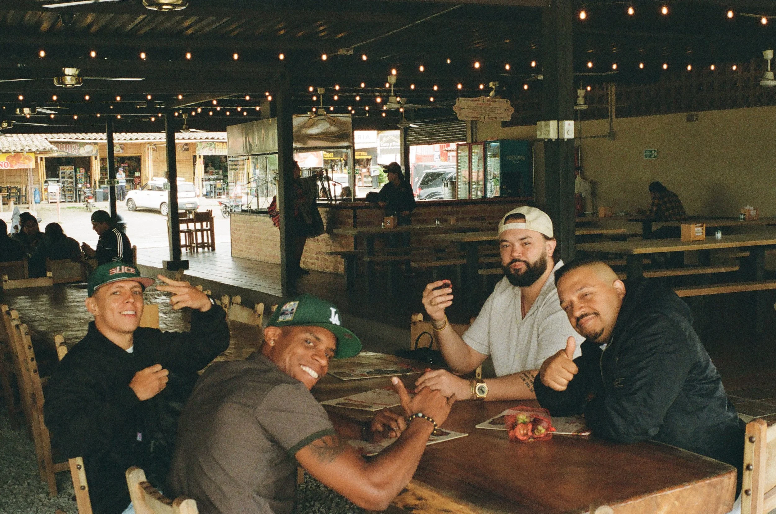 Four men sitting at a wooden table in a casual indoor restaurant or bar, smiling and posing for the camera with peace and thumbs-up gestures. The background shows a street view through open windows, with other patrons and string lights overhead.