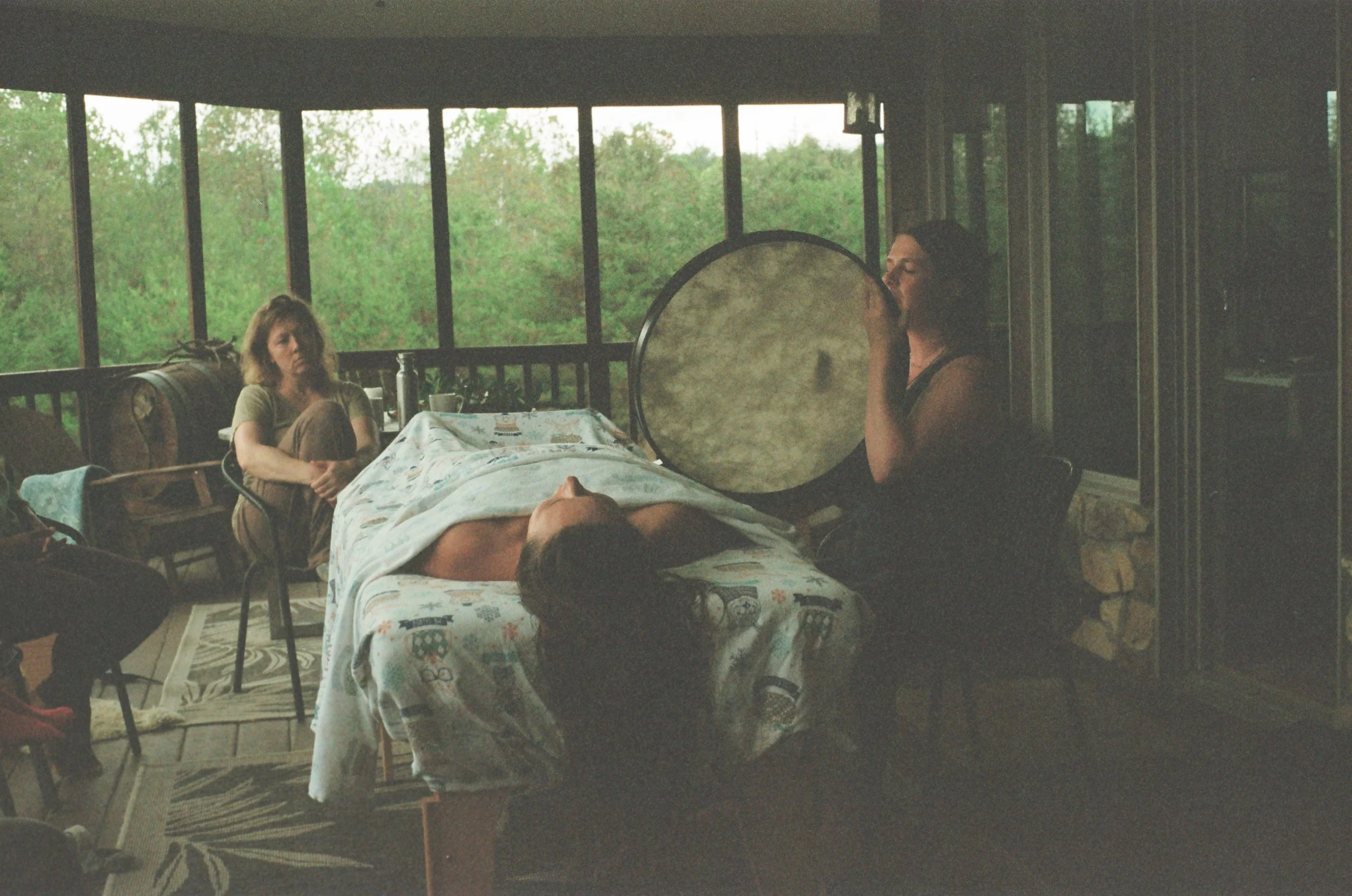 A woman lying on a bed with a floral blanket, appearing to be outdoors in a screened porch surrounded by trees, with two women sitting nearby, one playing a large frame drum.