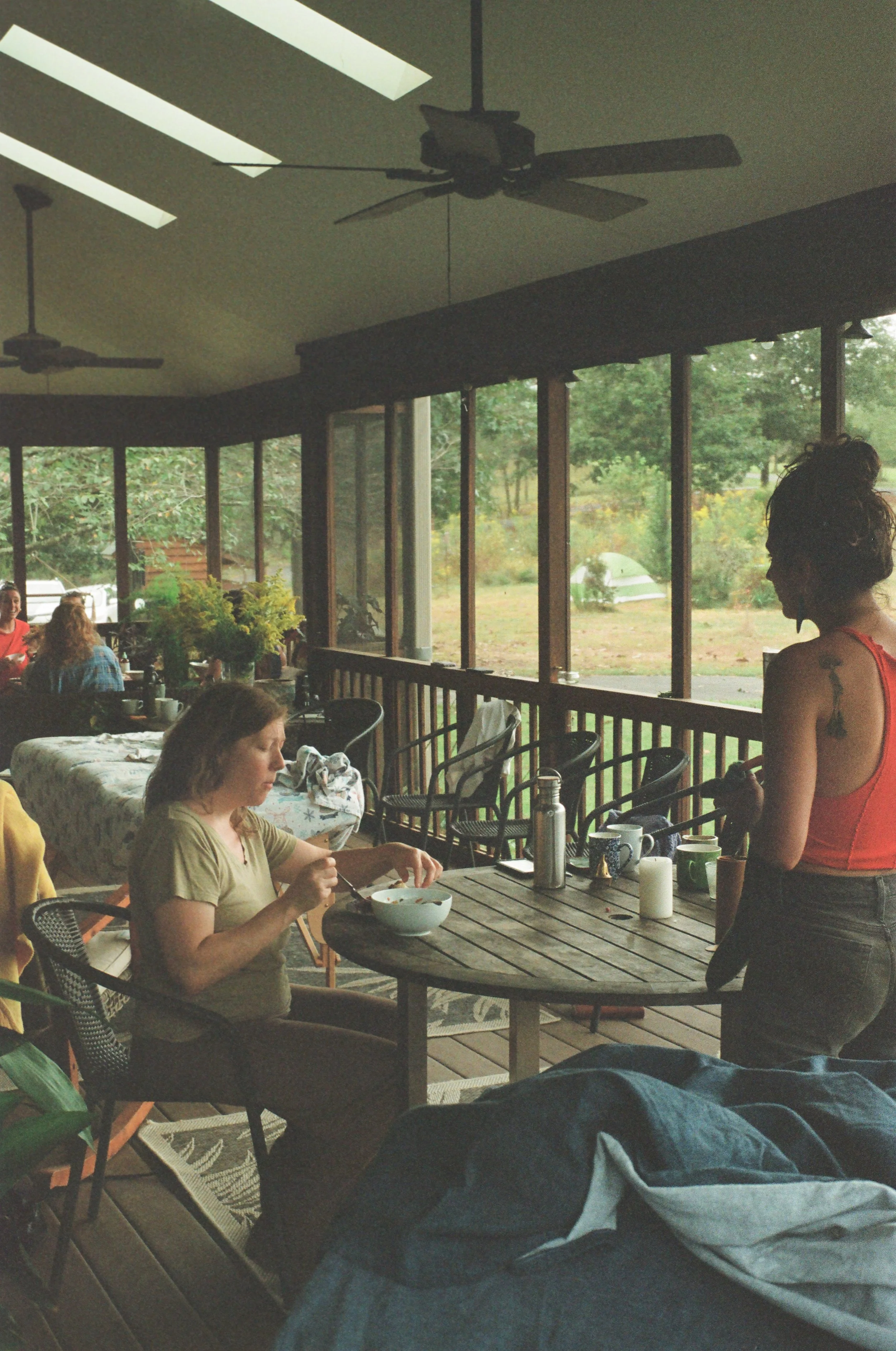 People inside a sunlit porch dining area with large windows overlooking greenery, some seated, a woman in a red top standing, and dining items on tables.