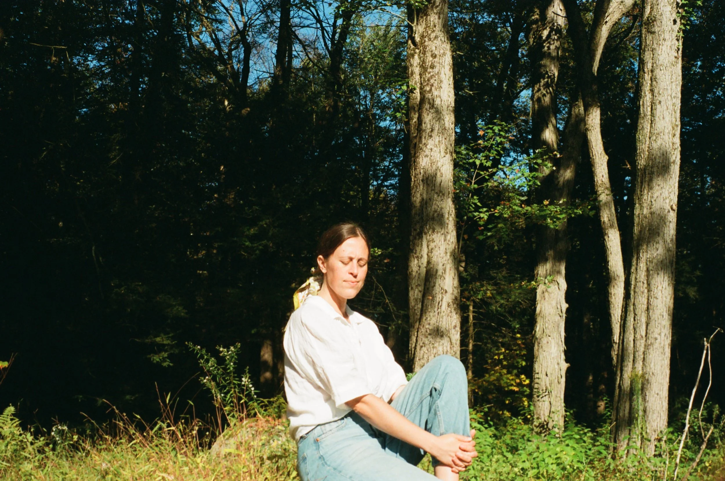 A woman with brown hair tied back in a ponytail, wearing a white shirt and jeans, sitting on the grass with her eyes closed in a forested area during daylight.