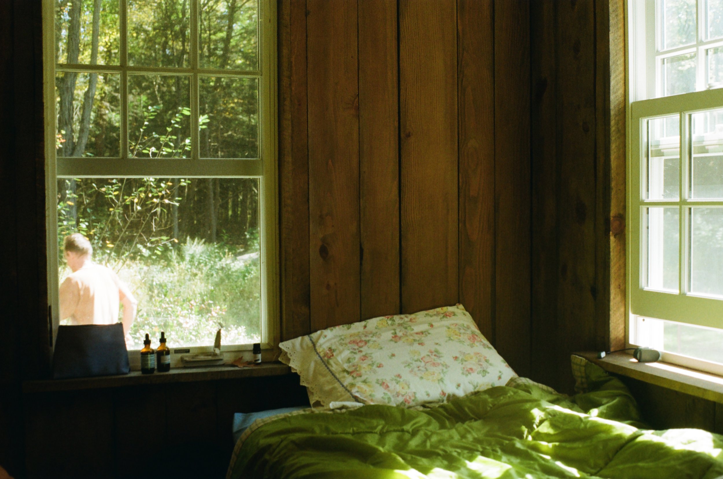 View of a bedroom with wooden wall panels, a bed with floral and green bedding, and two large windows showing greenery outside; a person is visible outside through the left window.