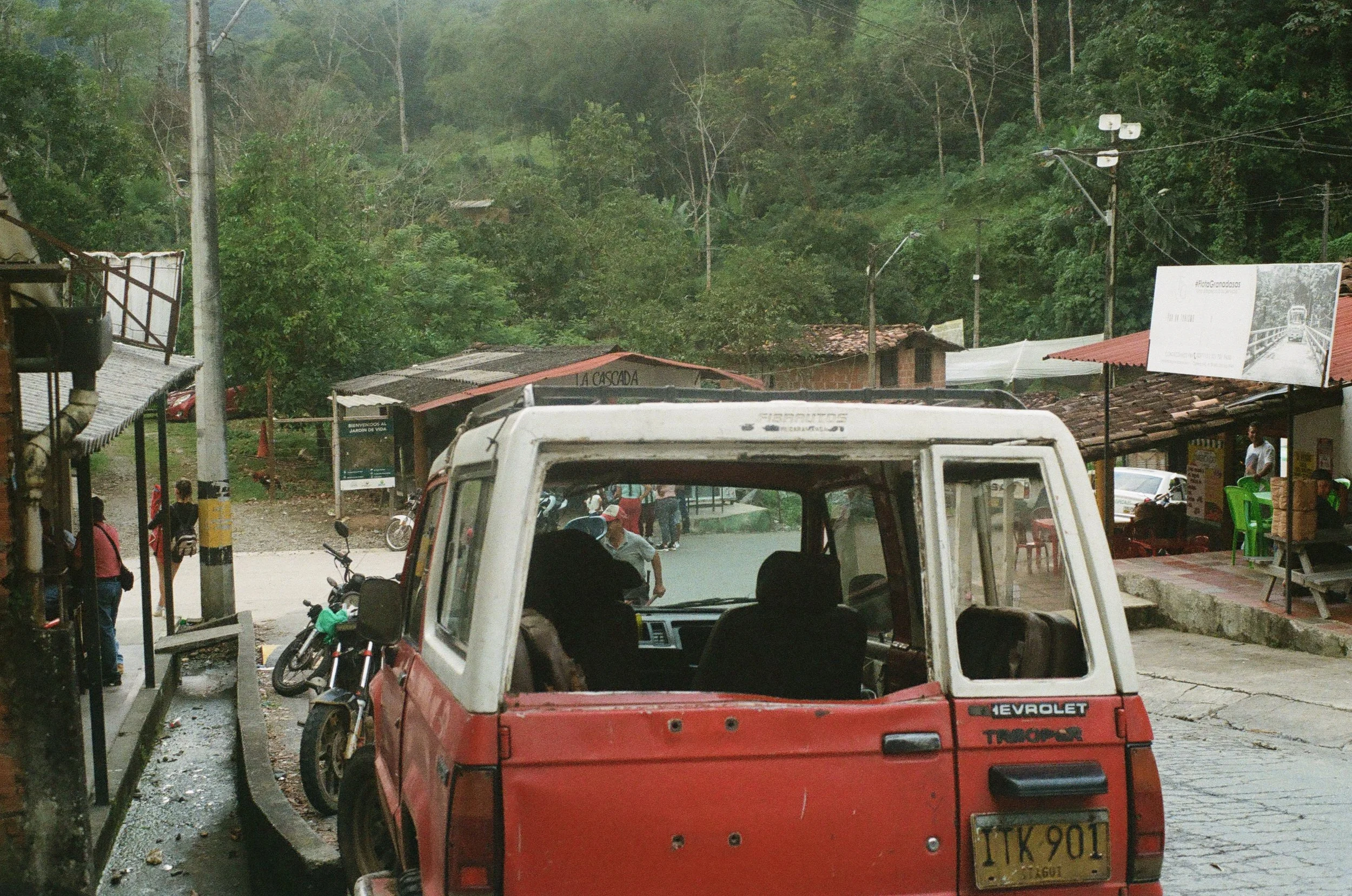 A red and white Chevrolet Trooper vehicle parked on a paved street in a small town with people walking around and a mountain with trees in the background.