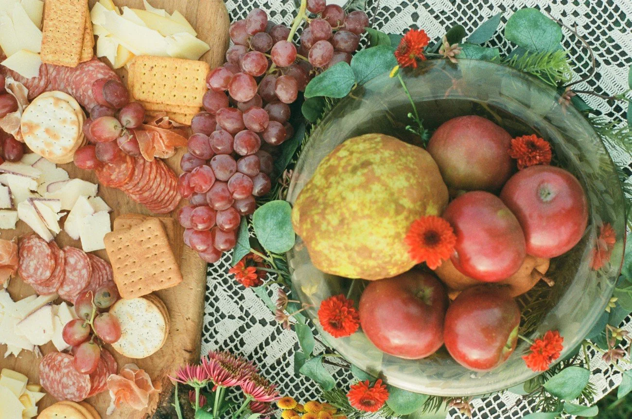 A charcuterie board with cheese, crackers, and cured meats, alongside a bunch of red grapes, and a glass bowl containing apples, a pear, and decorative red flowers on a table with lace tablecloth.