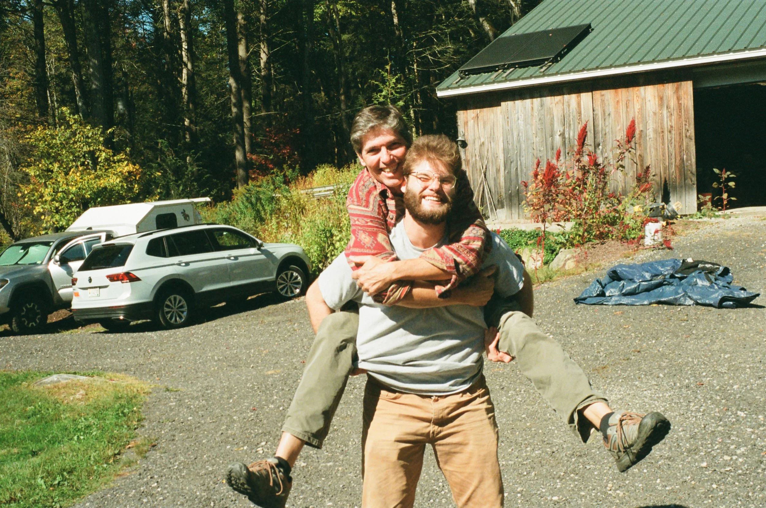 Two men outdoors, one carrying the other on his back, both smiling and happy, with a wooded background and parked cars nearby.