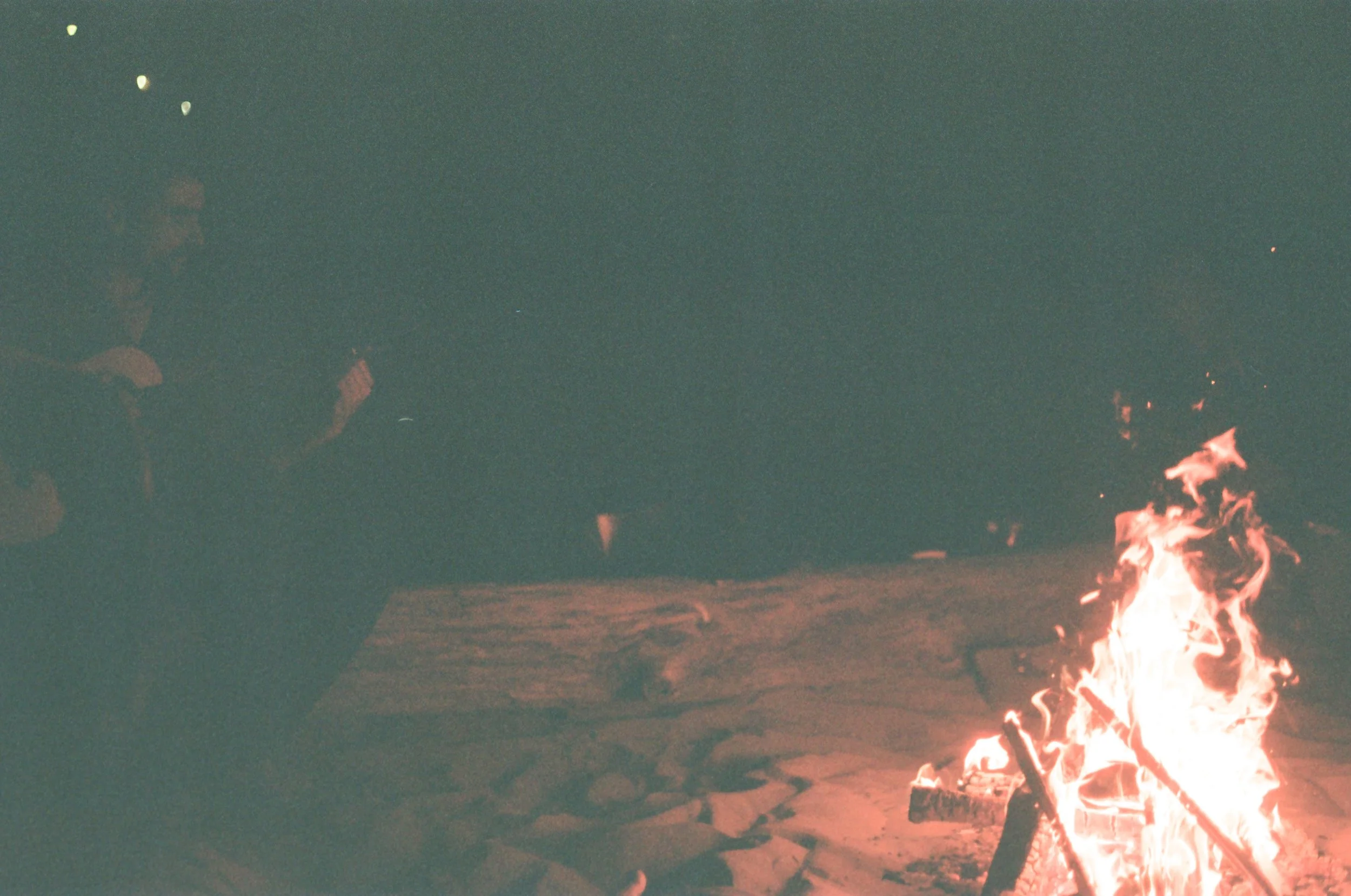 Nighttime scene of a campfire on a sandy surface with people gathered nearby, some holding drinks, with a dark sky overhead.
