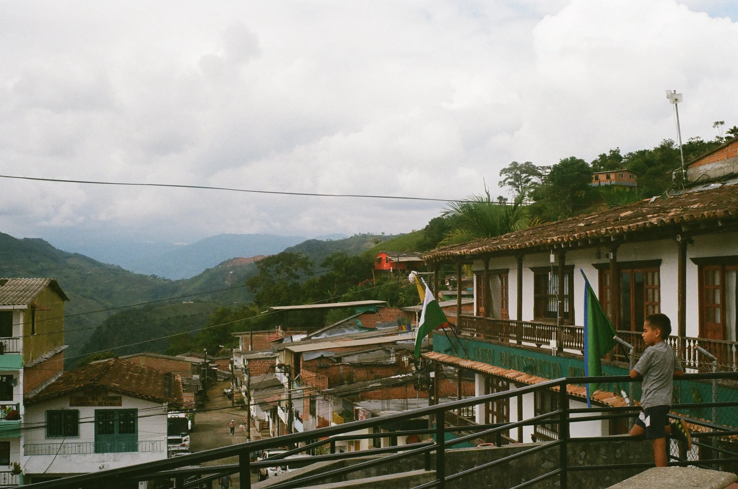 A view of a small town in the mountains with houses, flags, a boy standing on a terrace, and cloudy sky.