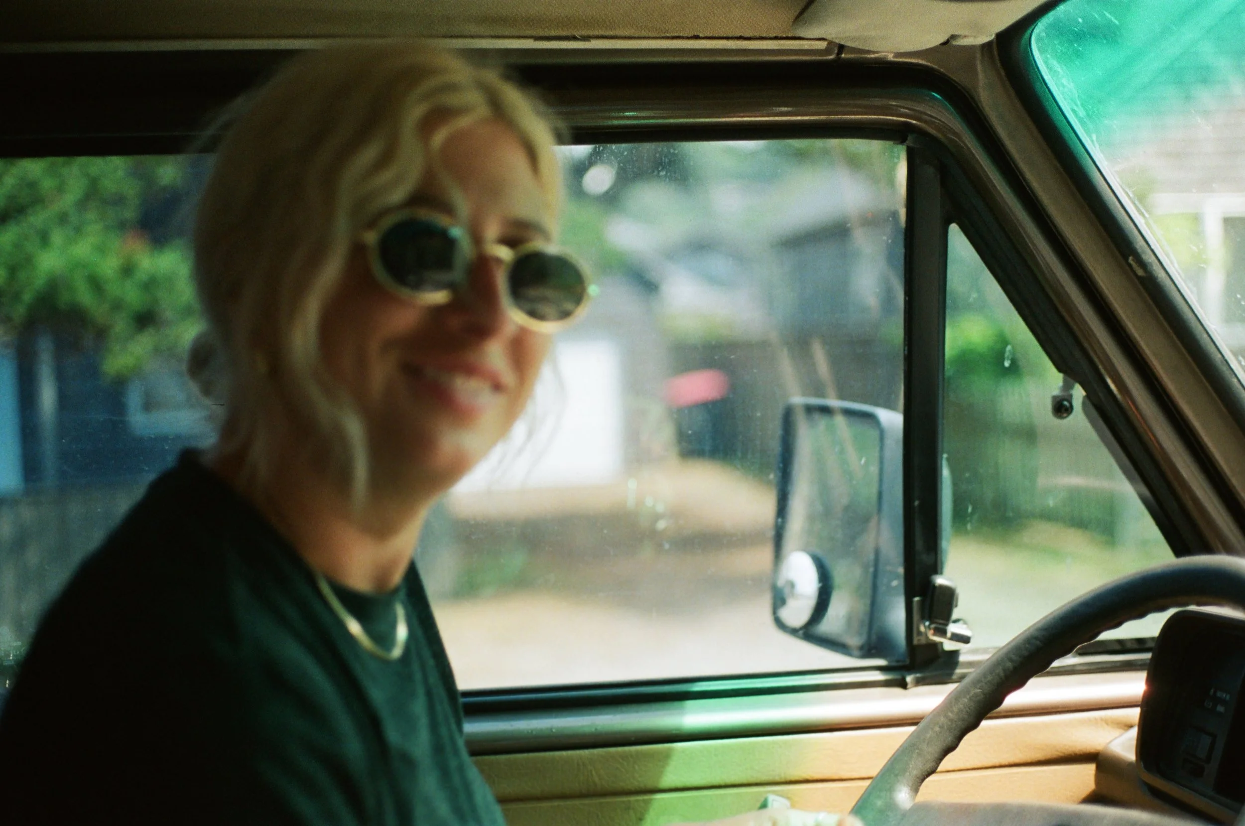 A woman with blonde wavy hair wearing round sunglasses, smiling inside a vehicle, with a view of greenery and a house outside through the window.
