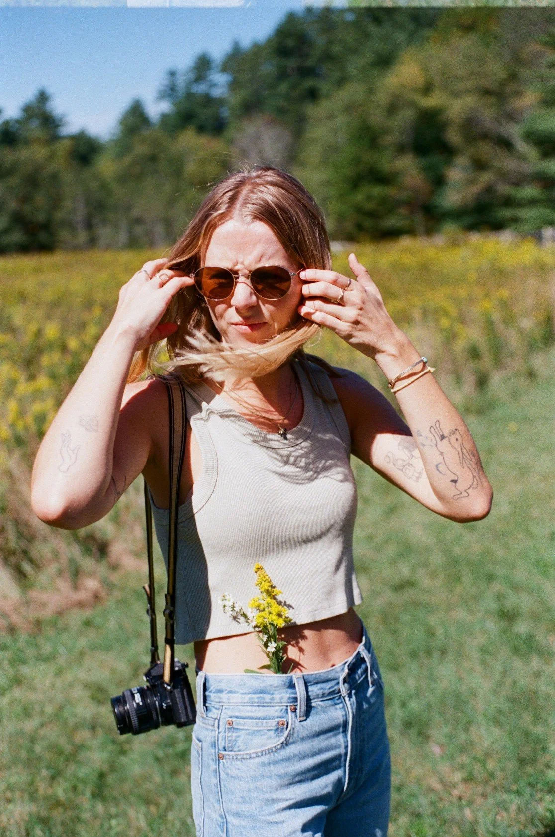 A young woman with long hair wearing sunglasses, a crop top, and jeans stands outdoors in a field with a camera hanging from her neck. She is adjusting her sunglasses with her hands, and a yellow flower is tucked into her jeans' waistband.