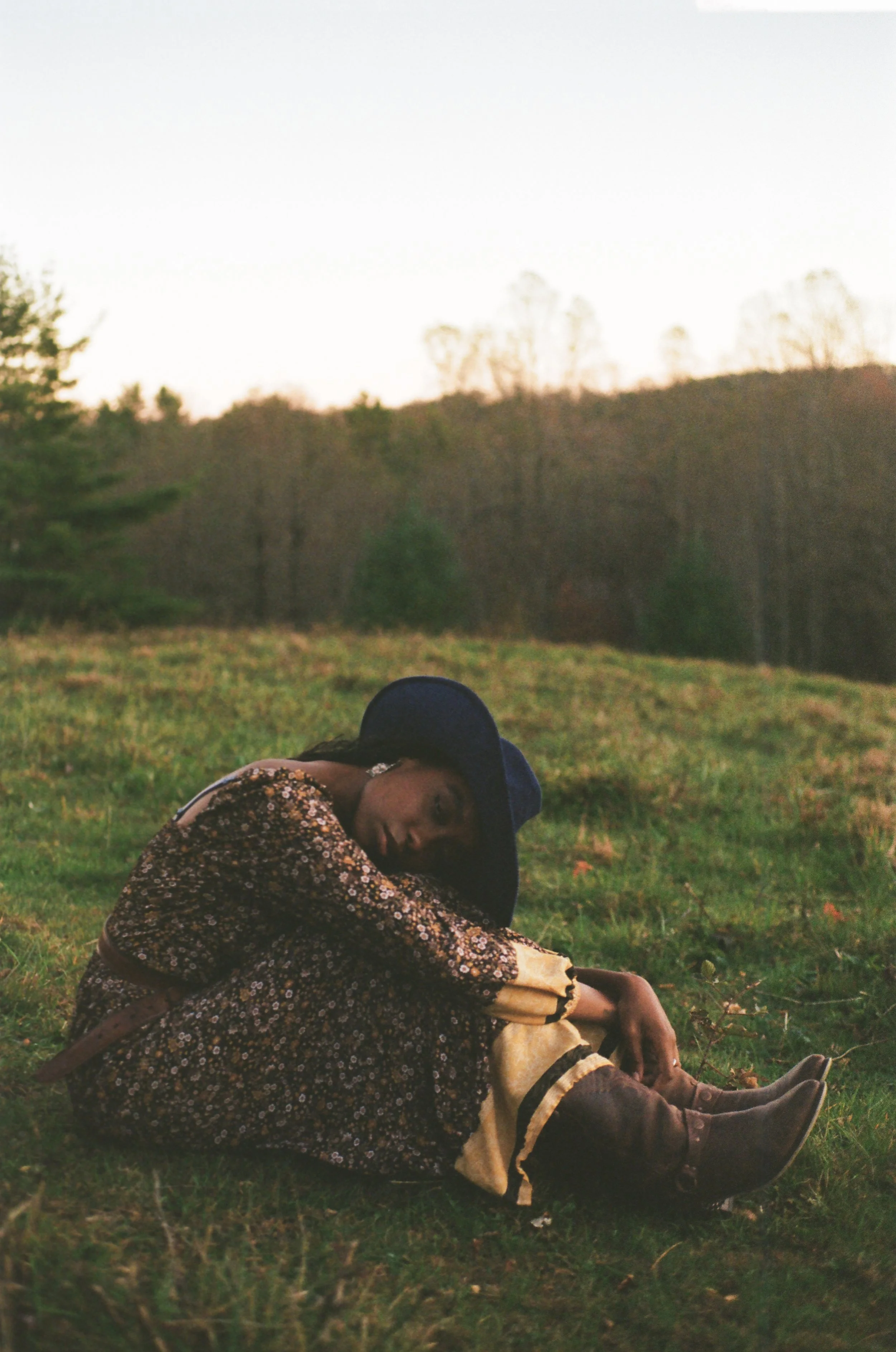 Woman sitting on grass in a field wearing a large blue hat and floral dress with brown boots, sunset or early evening sky in background.