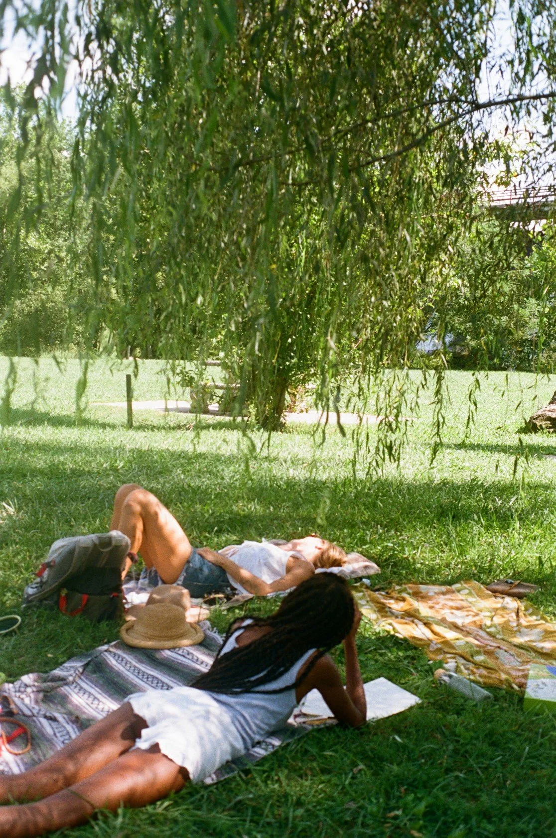 Two women relaxing outdoors in a park, one lying on her back on a blanket under a tree, and the other lying on her stomach, face down on a different blanket. There are hats, bags, and books nearby.
