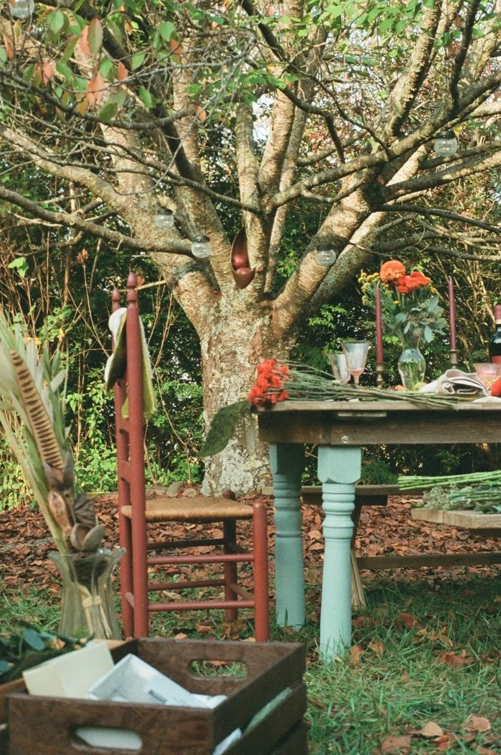 An outdoor memorial or altar setup in a garden, with a large tree in the background. The altar has flowers, candles, and various decorative items, with a chair and a table nearby.