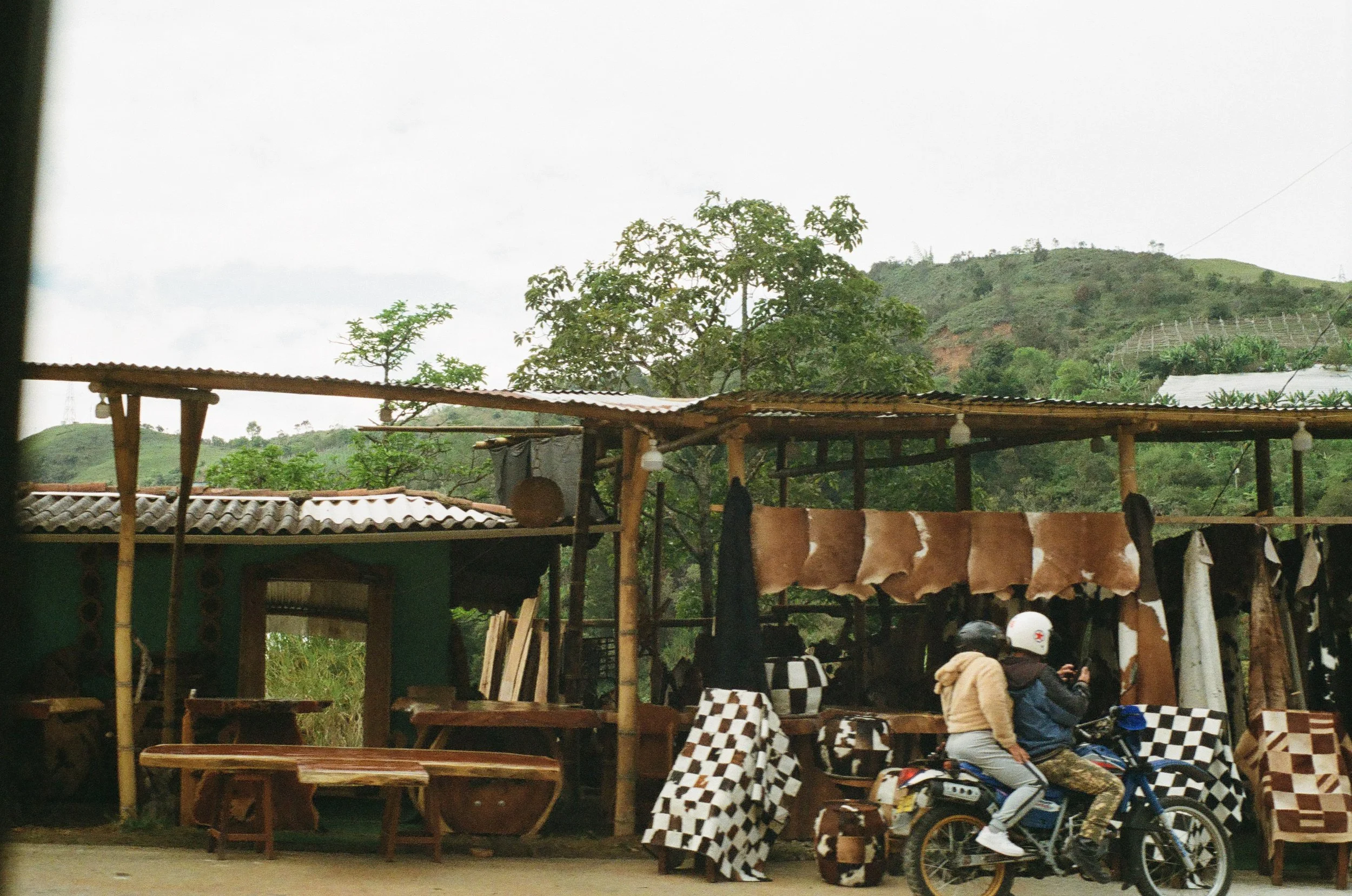 Two people riding a motorcycle past an outdoor market stall with animal hide and wood furniture, surrounded by trees and hills in the background.