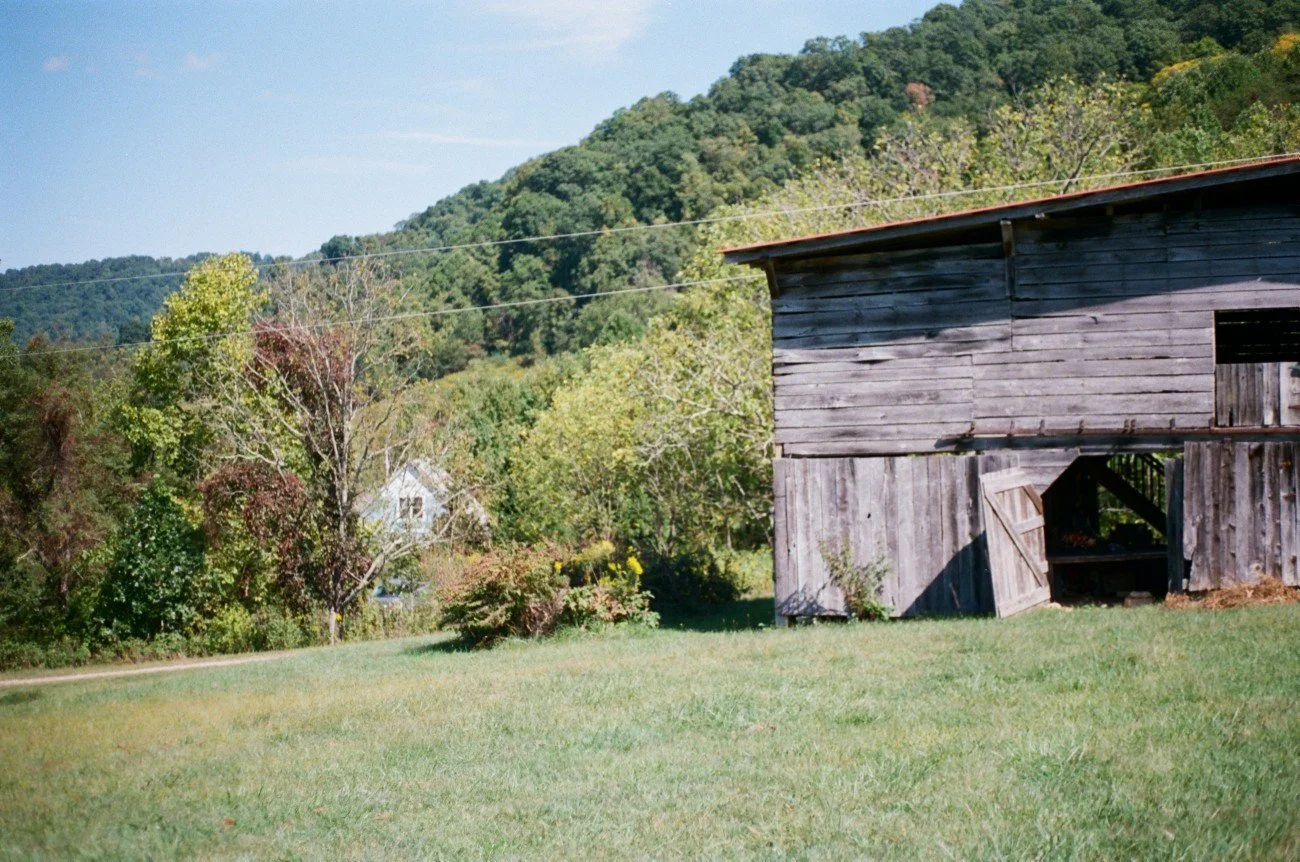 A rural scene with a wooden barn, grassy field, trees, and hills in the background.