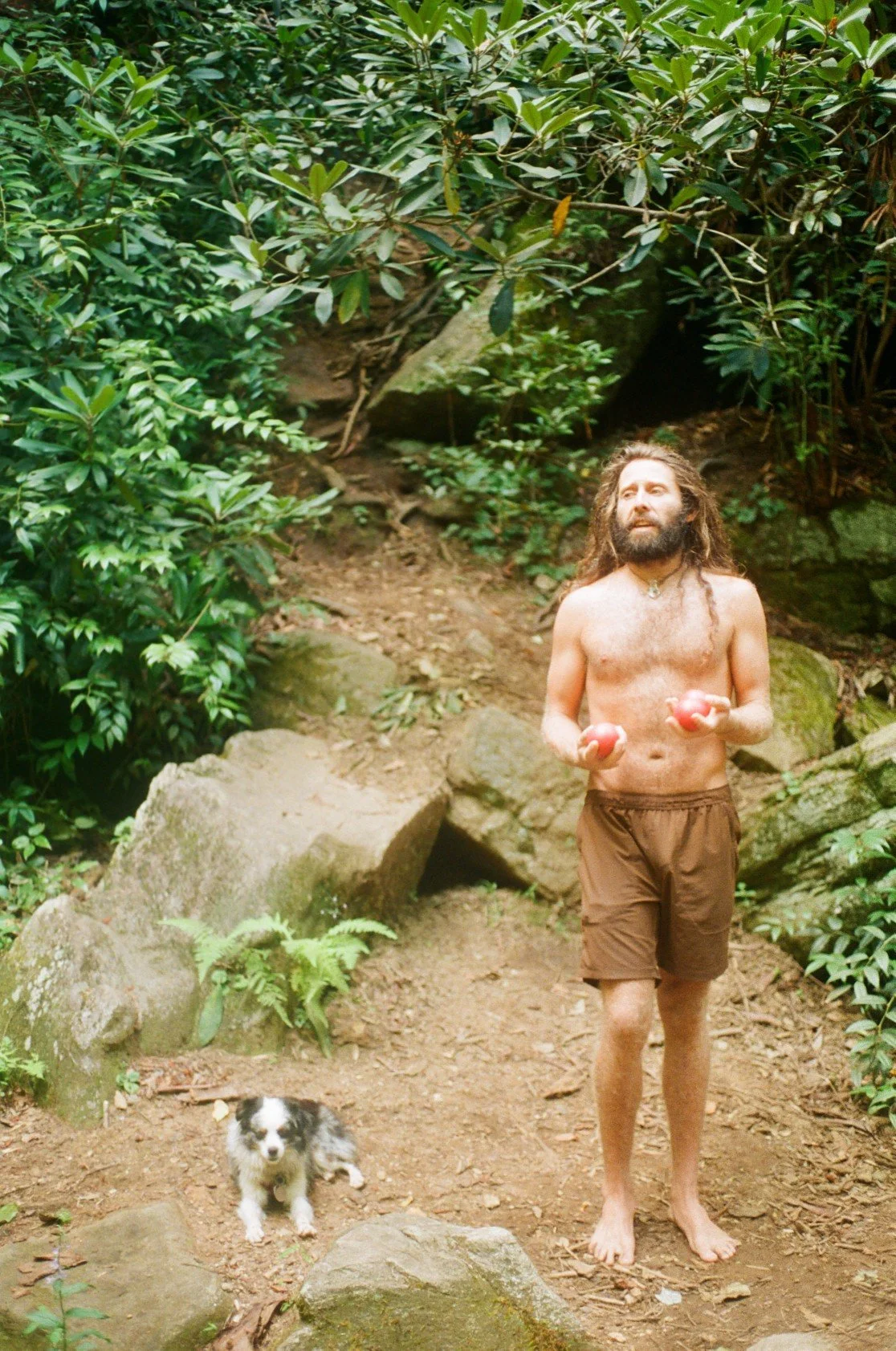 A shirtless man with long hair and a beard standing barefoot on a forest trail, holding two red balls, with a small black and white dog sitting nearby.