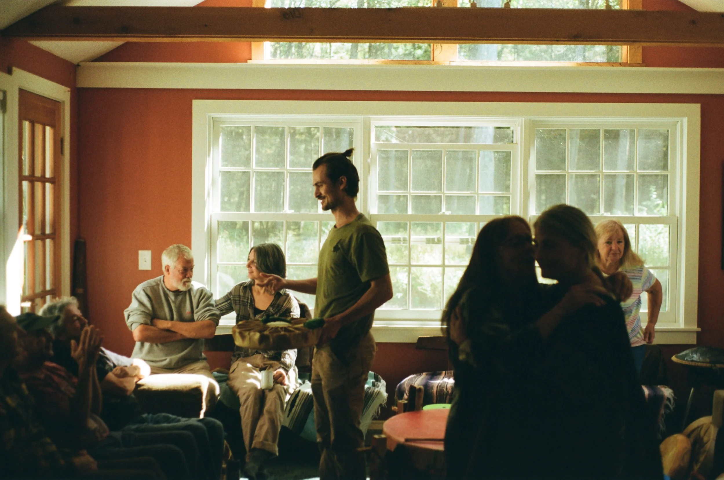 A group of people gathered in a well-lit room with large windows, some seated and some standing, engaged in conversation and laughter at what appears to be a social gathering or family reunion.