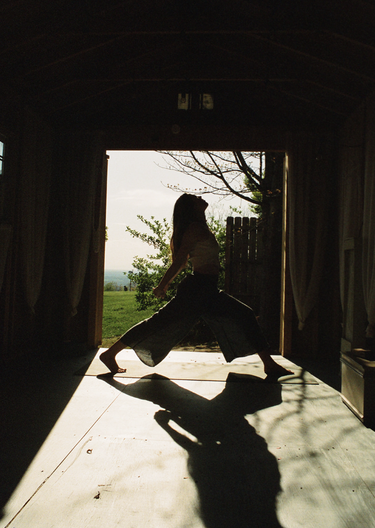 Silhouette of a person practicing yoga or dance in a barn opening with outdoor trees and sky visible in the background.