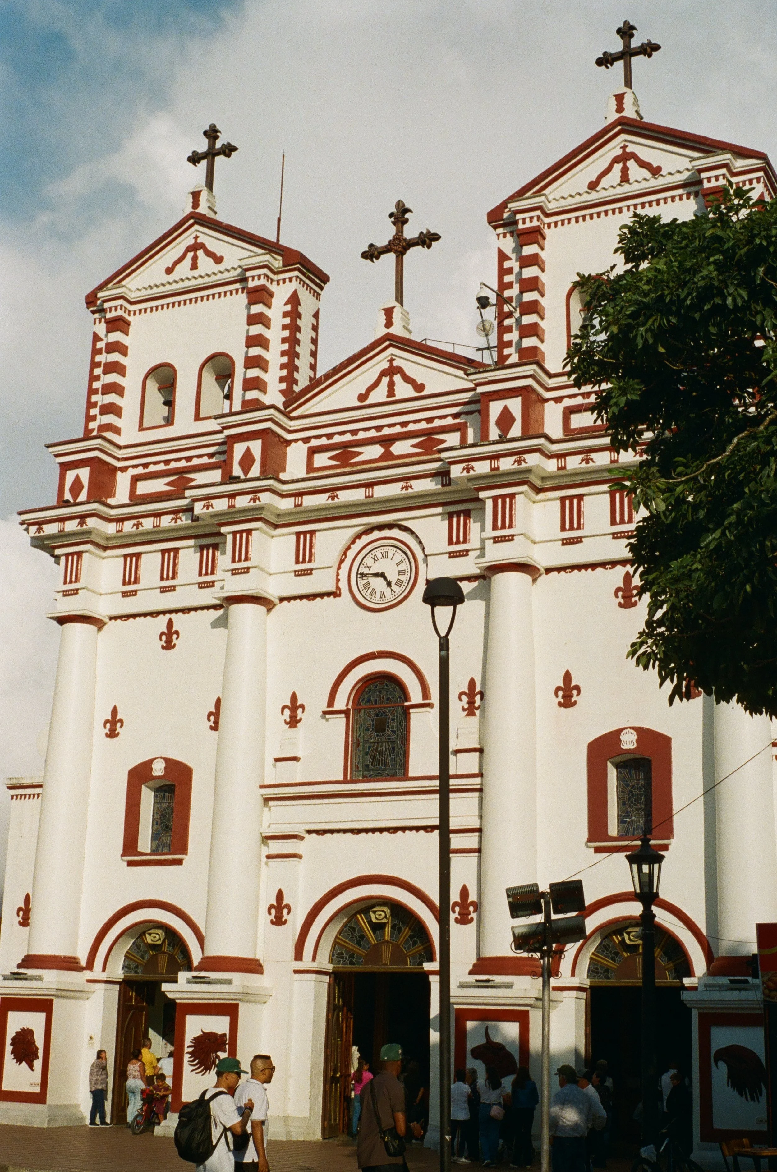 A white church with red accents and two bell towers topped with crosses, with a clock in the center and stained glass windows, with people gathered outside.