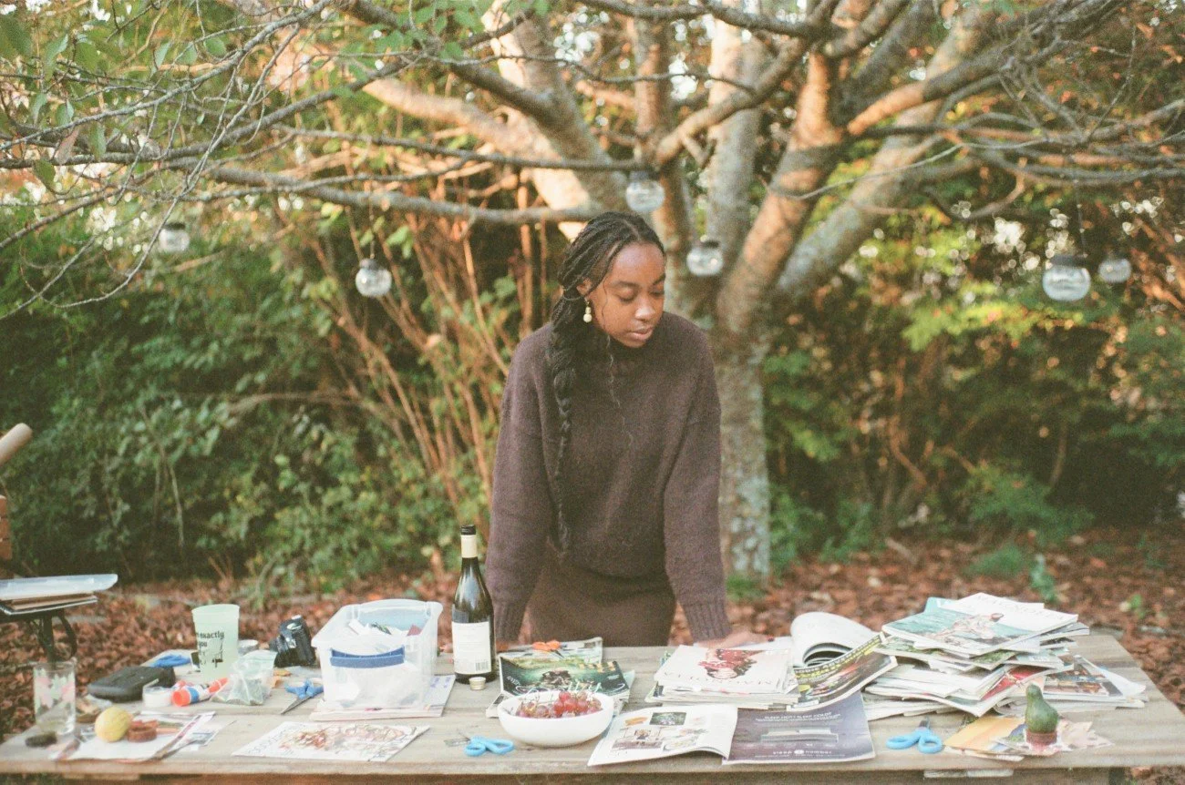 A woman with braids and earrings standing behind a table outdoors surrounded by trees, with magazines, papers, a bottle, a bowl, and scissors on the table.