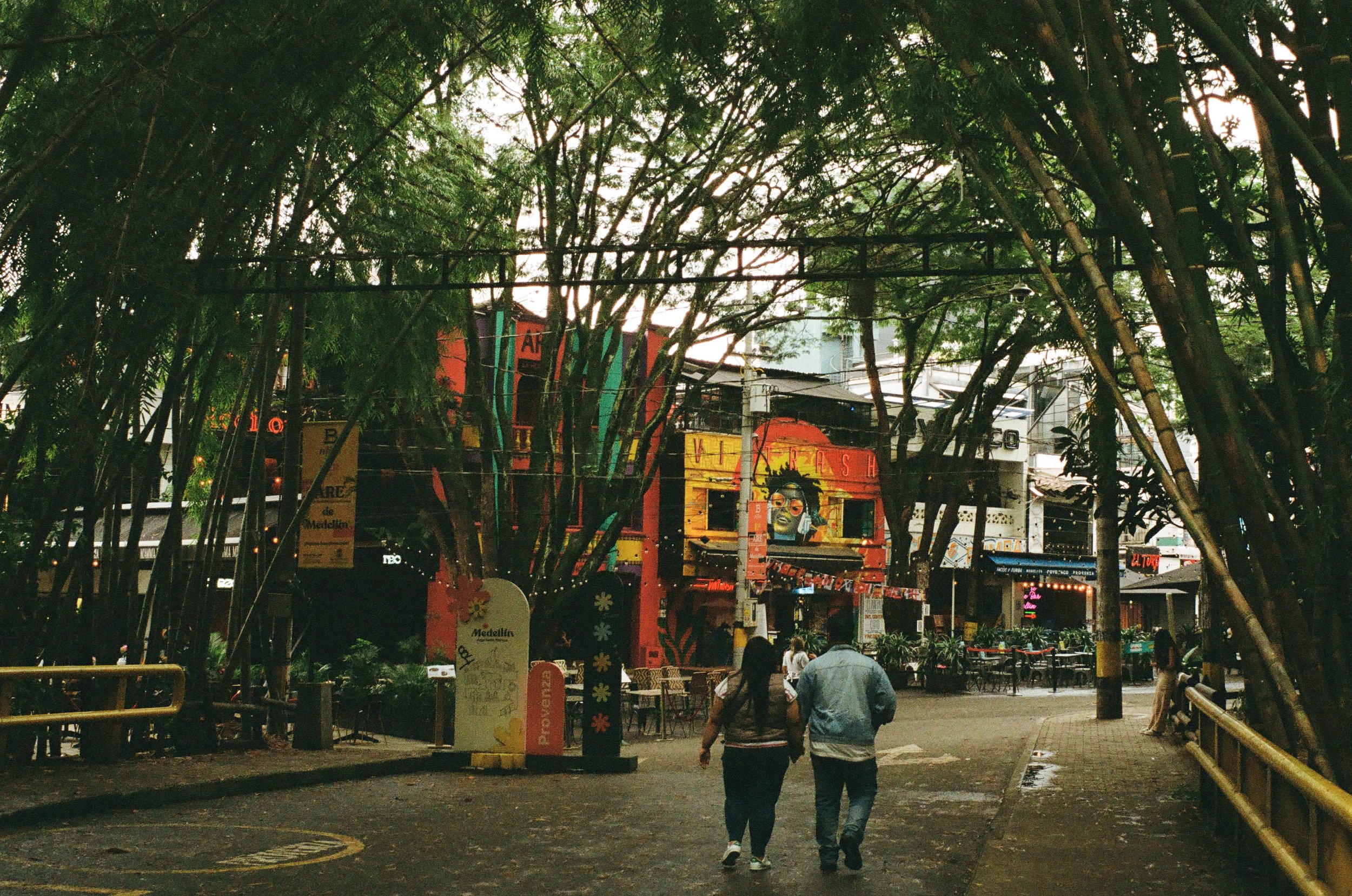 Two people walking down a street with trees and colorful buildings, outdoor seating, and signs, likely in a city area.