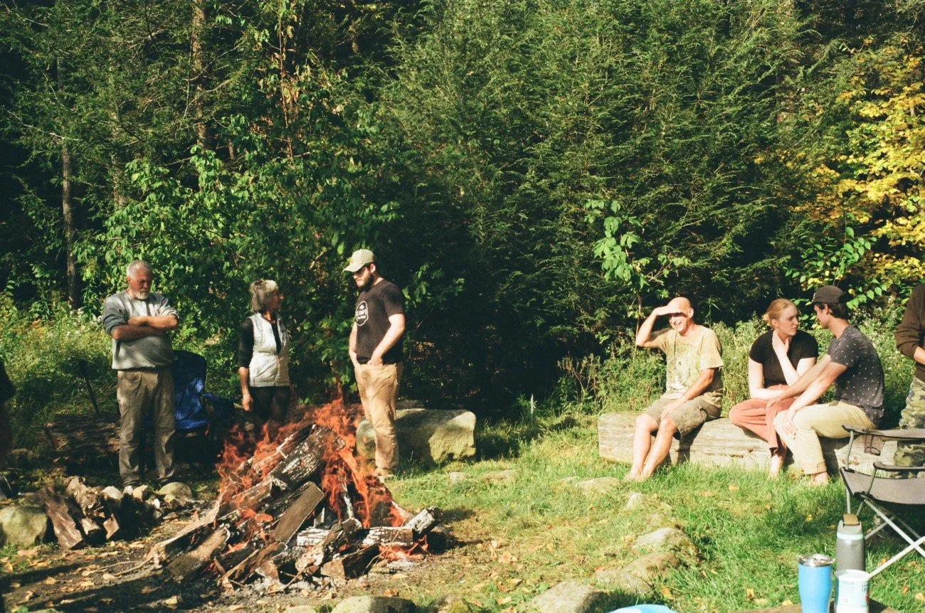 Group of people around a campfire in a forest, with some sitting on logs and others standing or sitting nearby.