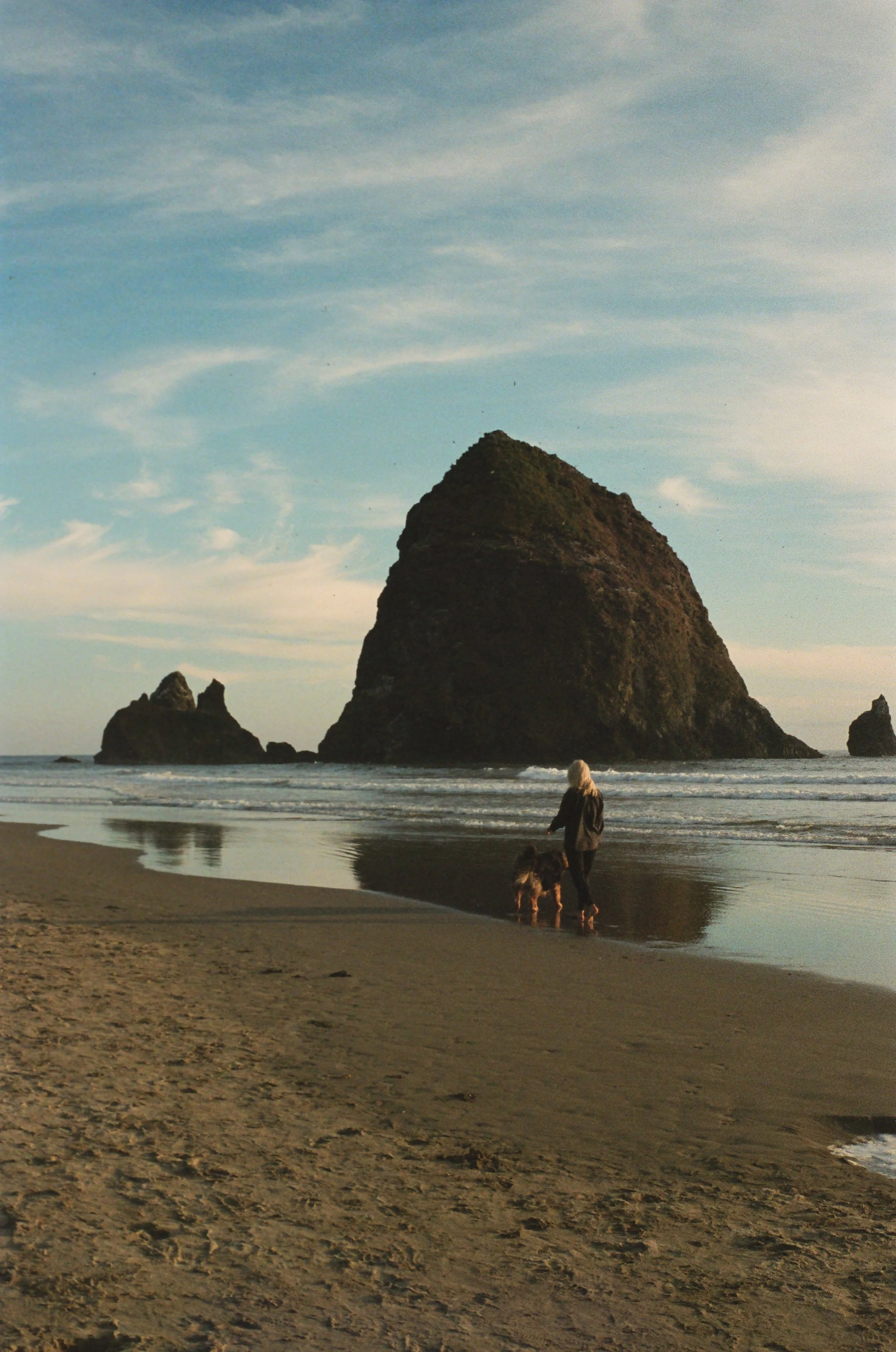 A person walking a dog on a beach with large sea stacks in the background, sand and water in the foreground, and a partly cloudy sky.