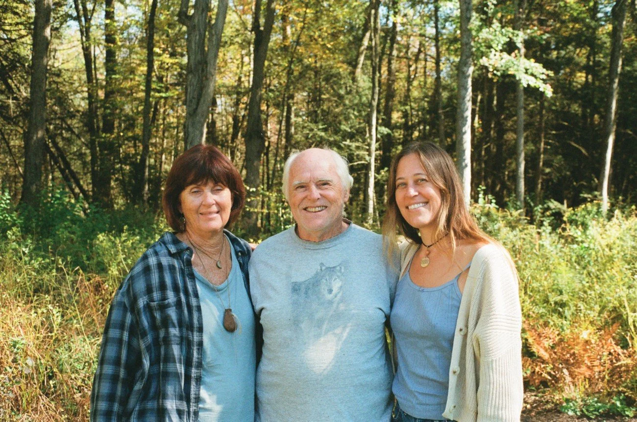 Three people smiling outdoors in a forest with green foliage and trees in the background.