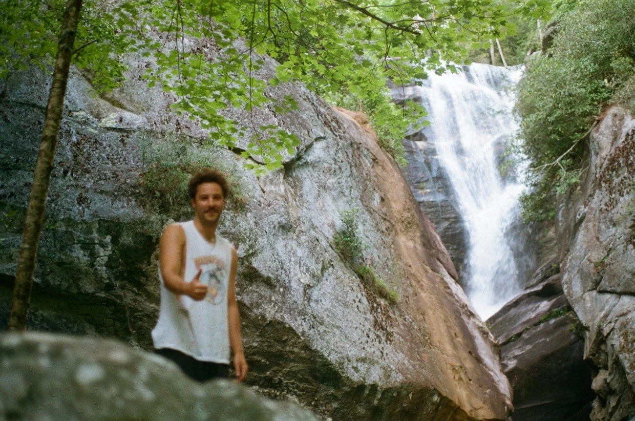 A man in a sleeveless white shirt smiling and giving a thumbs-up, standing in front of a waterfall surrounded by rocks and lush green trees.